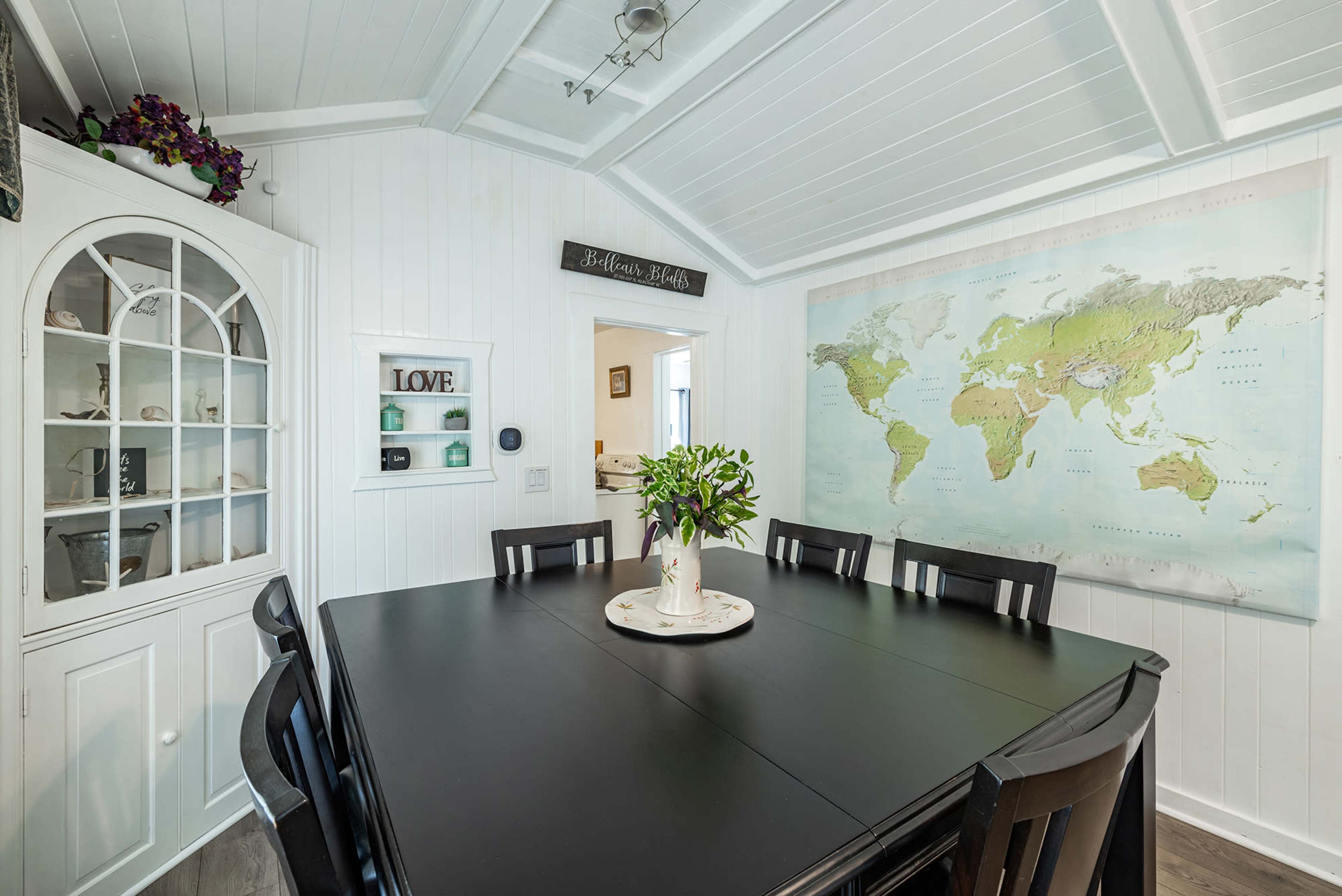 A dining area features a large black table surrounded by chairs, with a world map hung on the wall and a glass cabinet displaying decorative items.