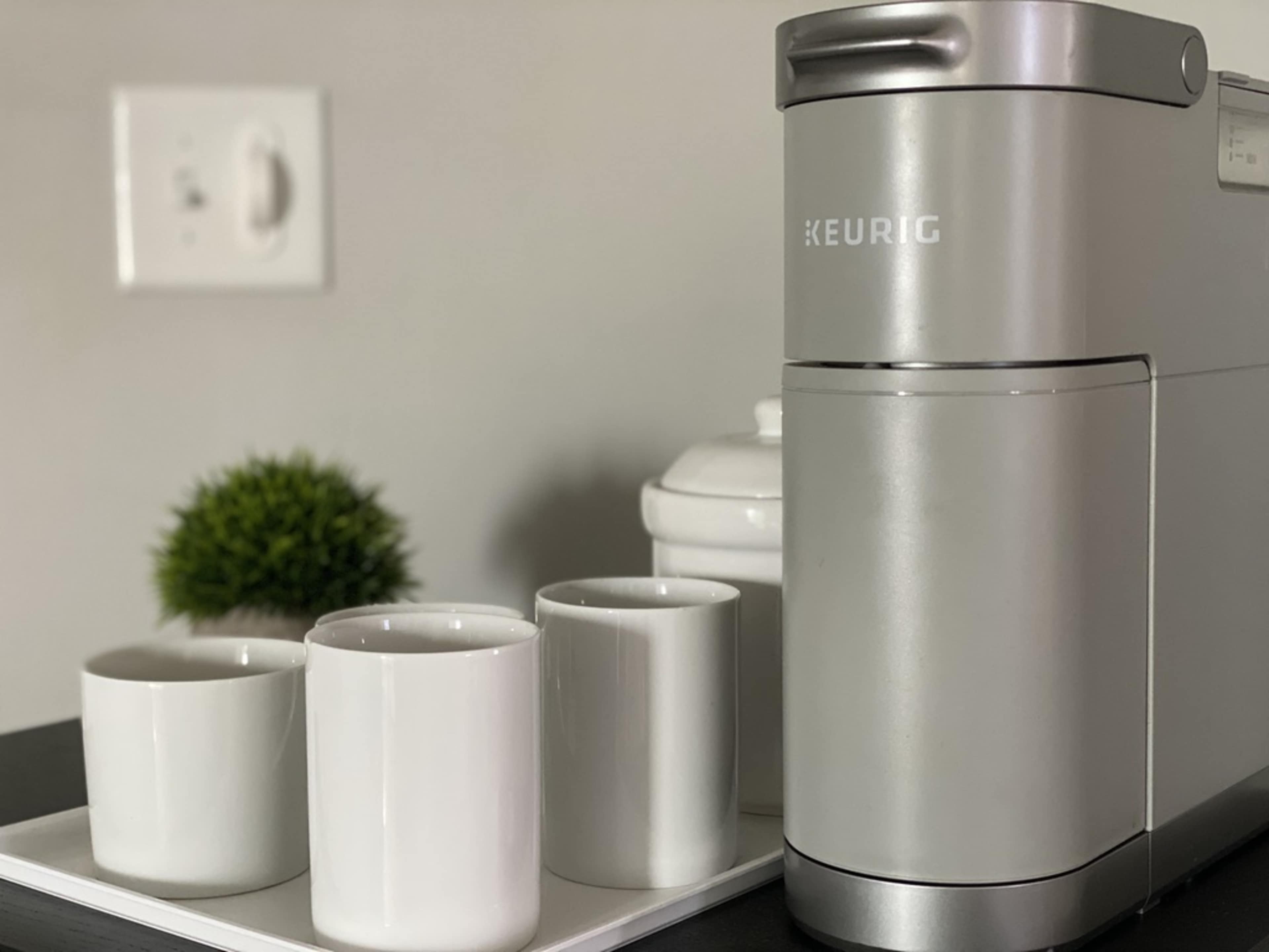 A silver Keurig coffee maker sits next to three white coffee mugs on a tray, with a small green plant in the background.