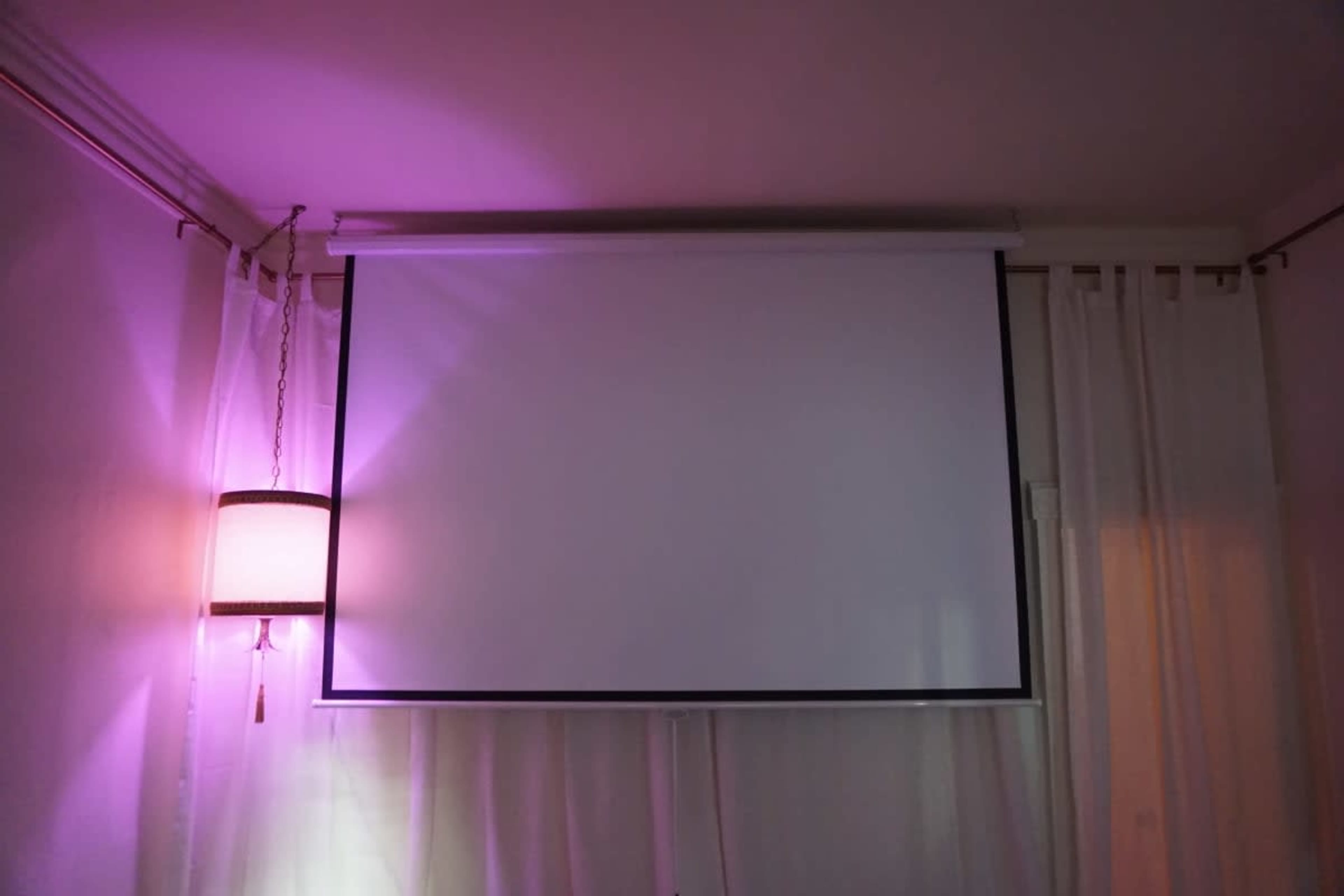 A white projection screen is mounted on a wall beside a lamp with a purple glow.