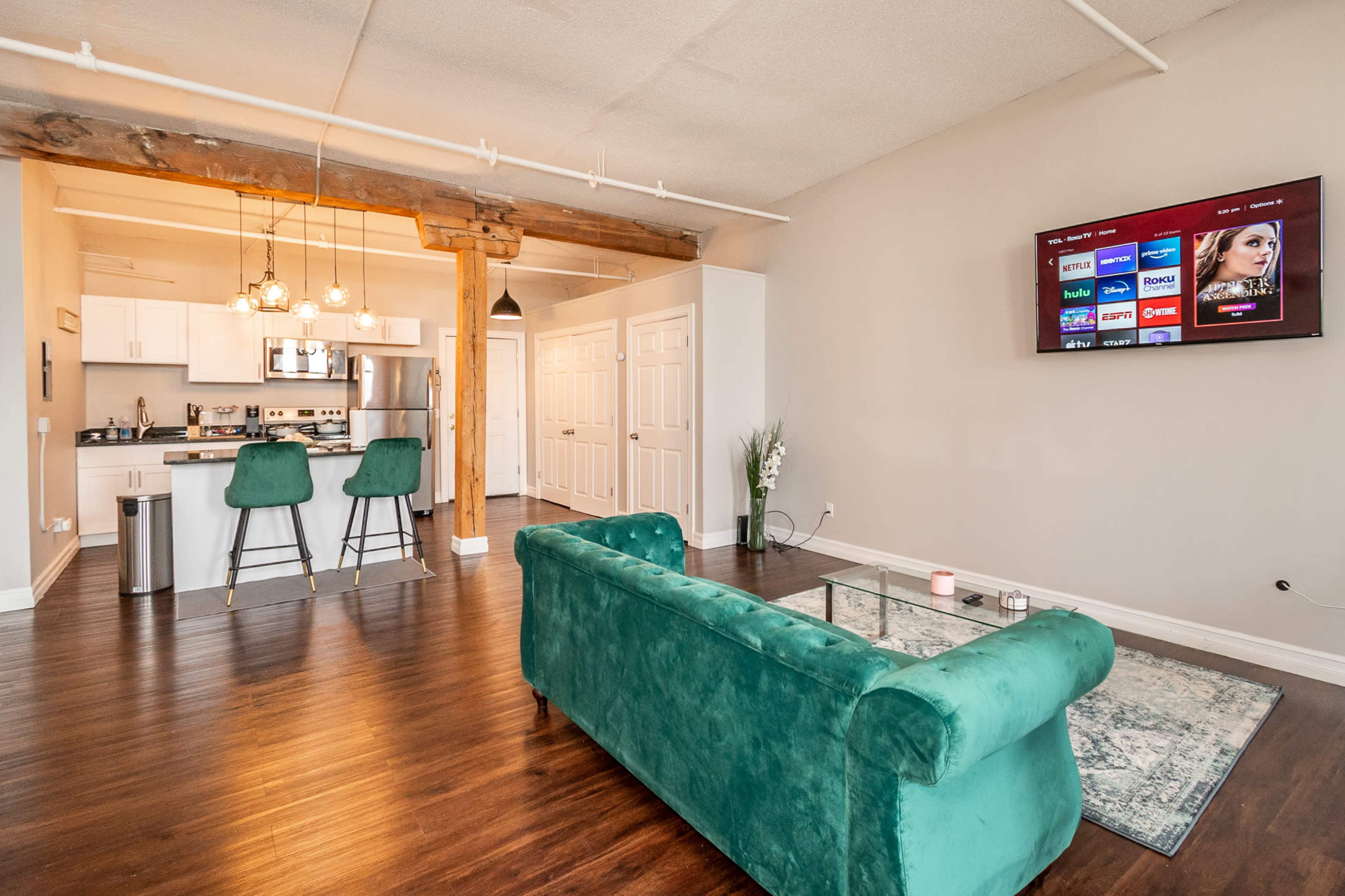 A modern living area with a green sofa, a kitchen bar with two stools, and a wall-mounted TV.