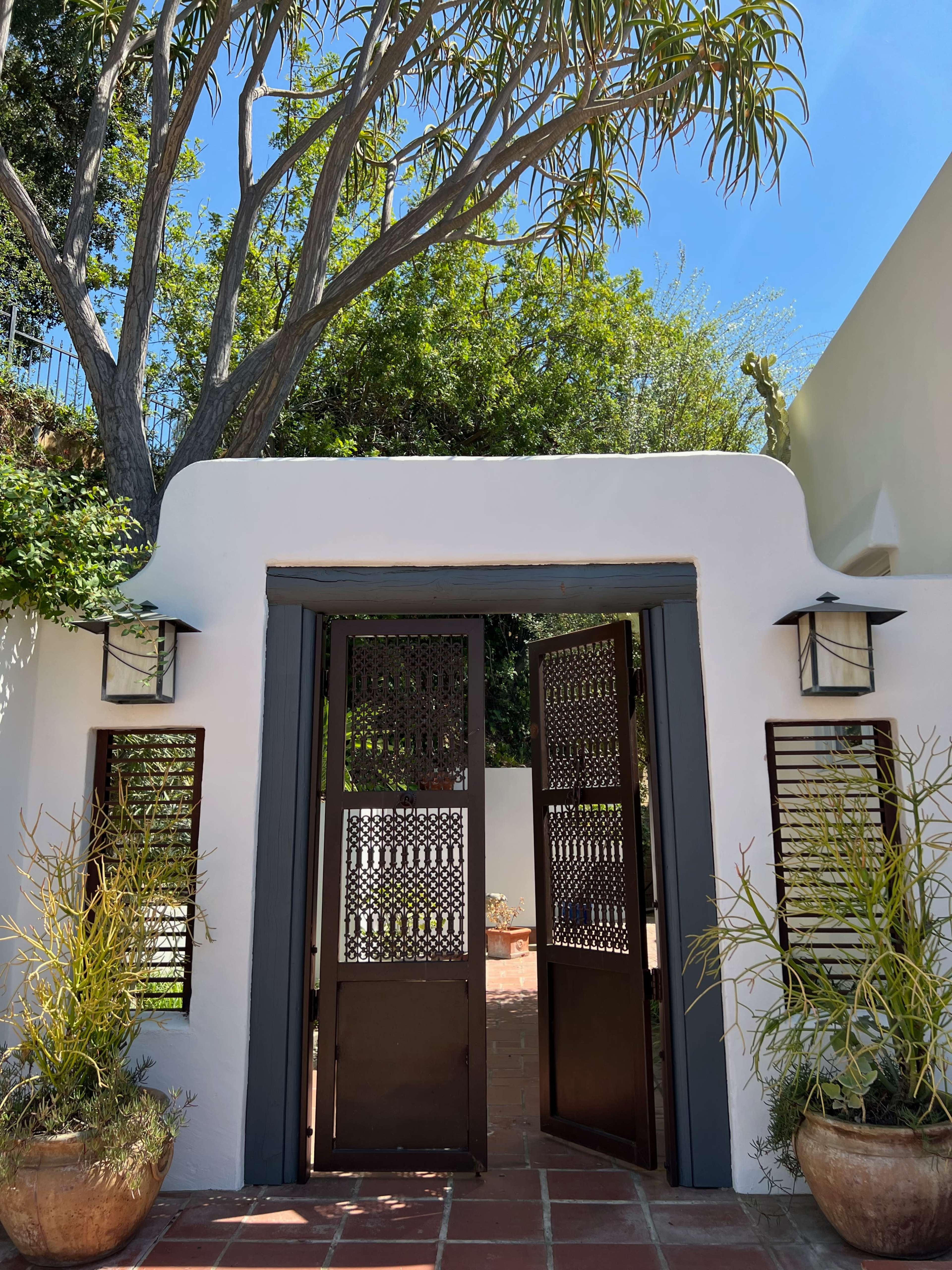 A pair of wooden doors open into a courtyard surrounded by greenery, with a tree providing shade above.