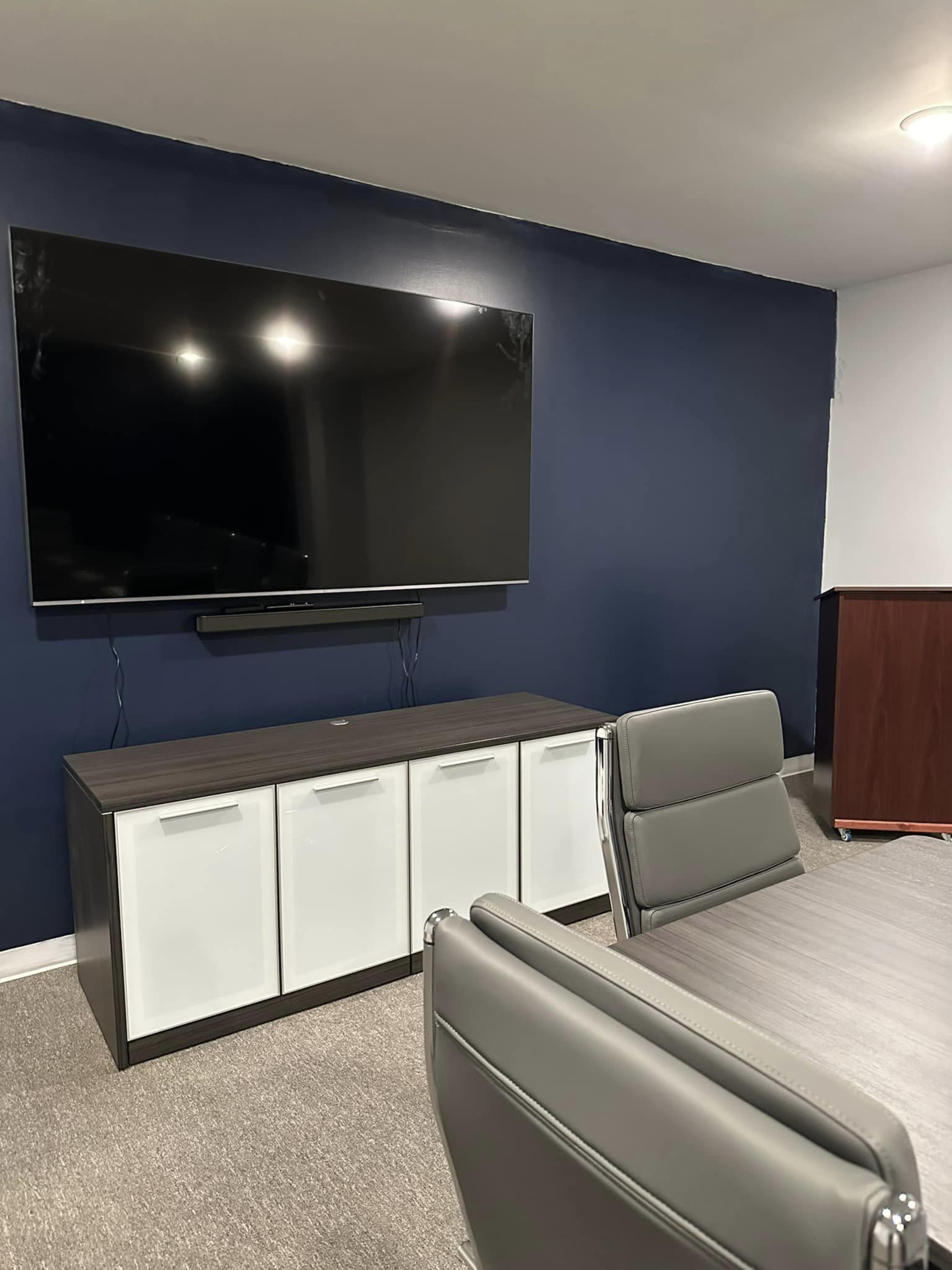 A modern office space features a large flat-screen TV mounted on a dark blue wall above a sleek cabinet with white and dark wood doors, accompanied by a conference table and chairs.
