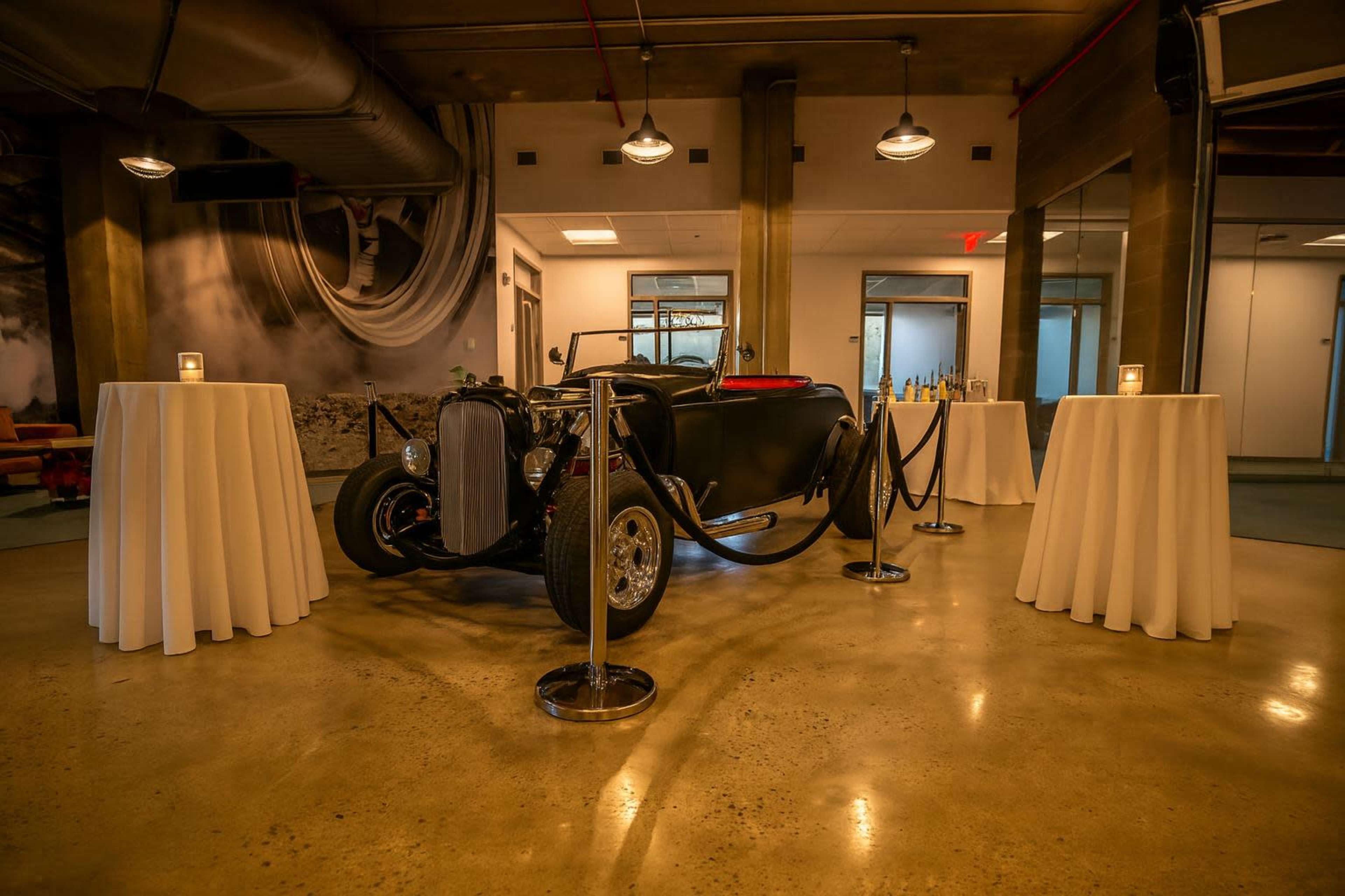 A vintage car is displayed in the center of a spacious room, surrounded by round tables draped in white fabric and delineated by black rope barriers.