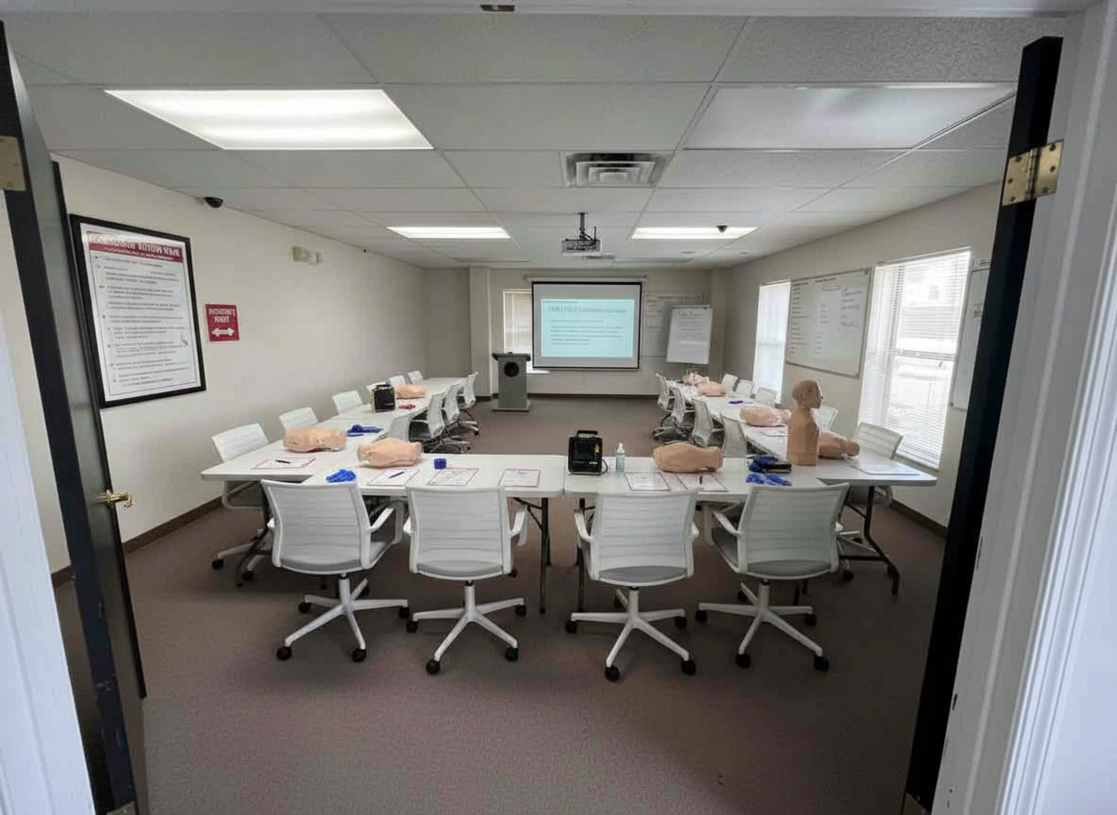 A classroom is set up with white chairs arranged around a long table, featuring training mannequins and presentation equipment in front of a screen.