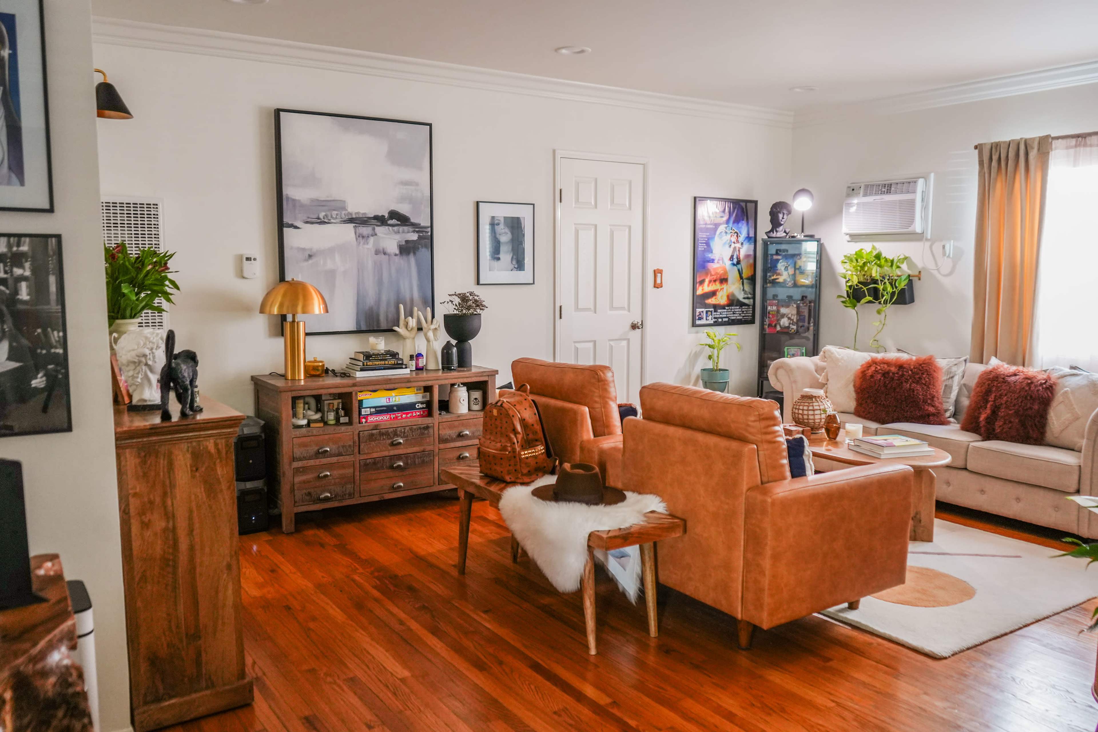 The image shows a cozy living room with brown leather furniture, a wooden cabinet, and various artworks on the walls.