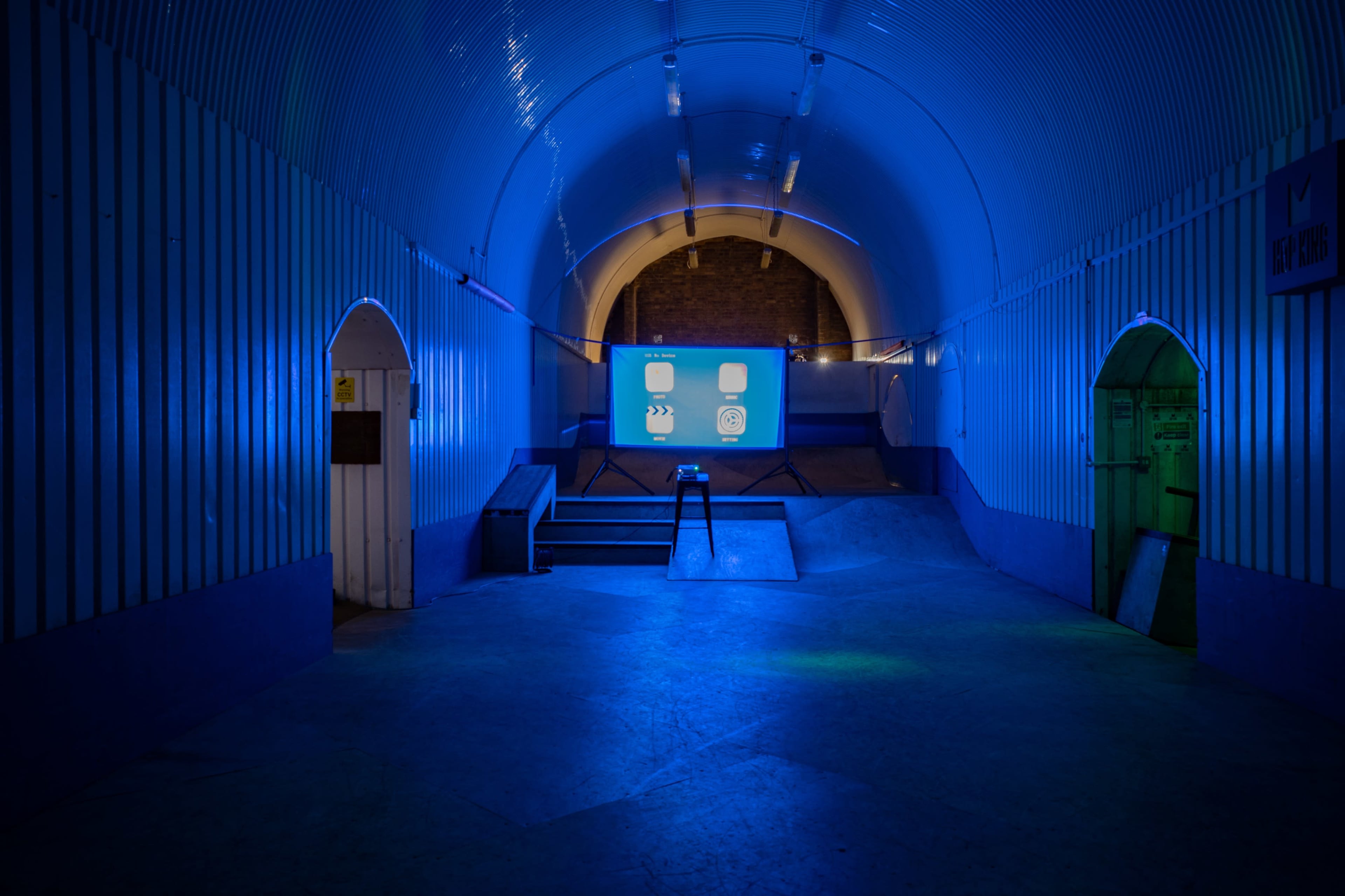 The scene shows a dimly lit tunnel with blue lighting, featuring a small stage at the center and a projection screen at the far end.