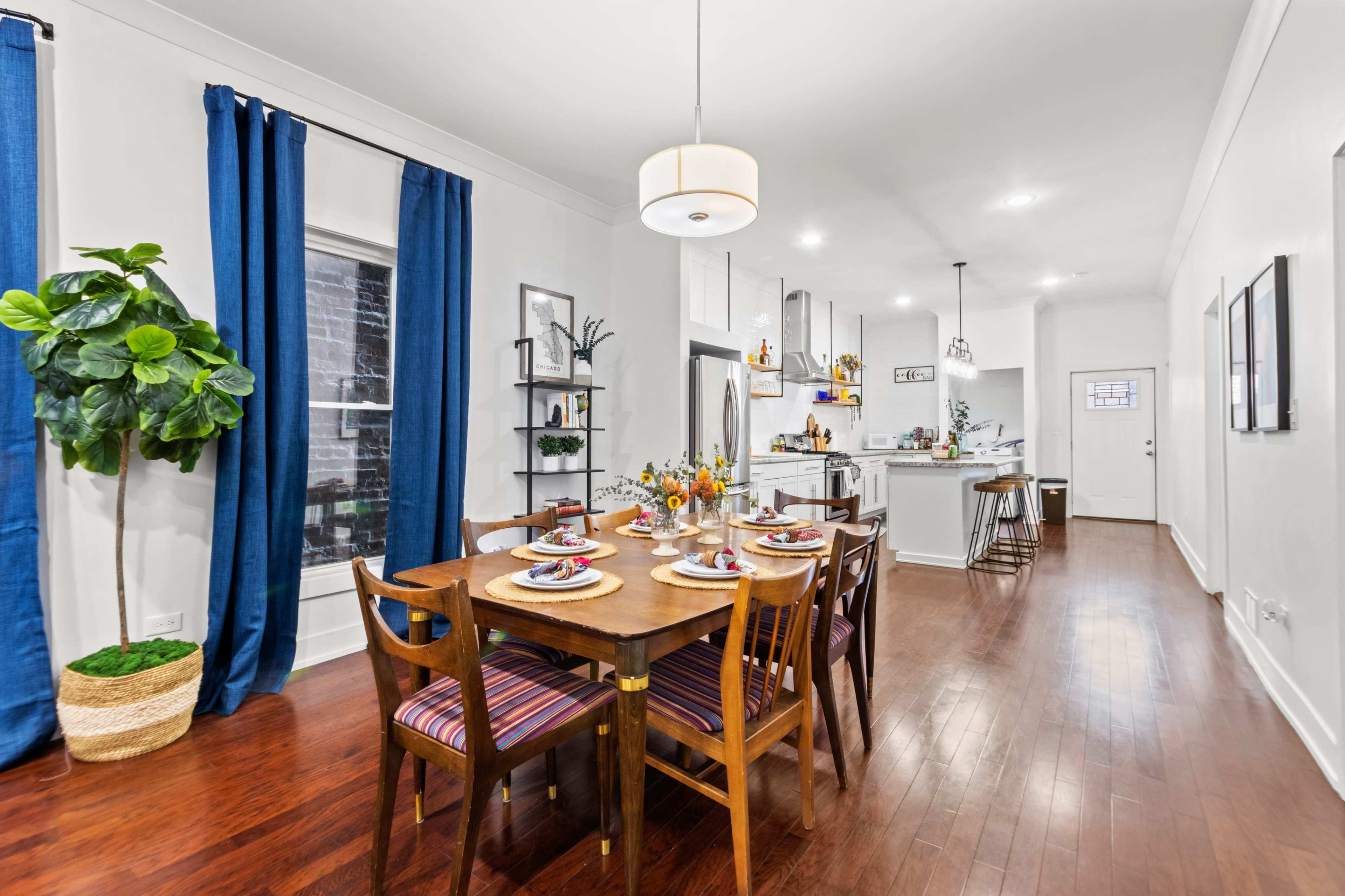A dining area with a wooden table set for a meal, surrounded by chairs, with blue curtains, a potted plant, and an open kitchen in the background.