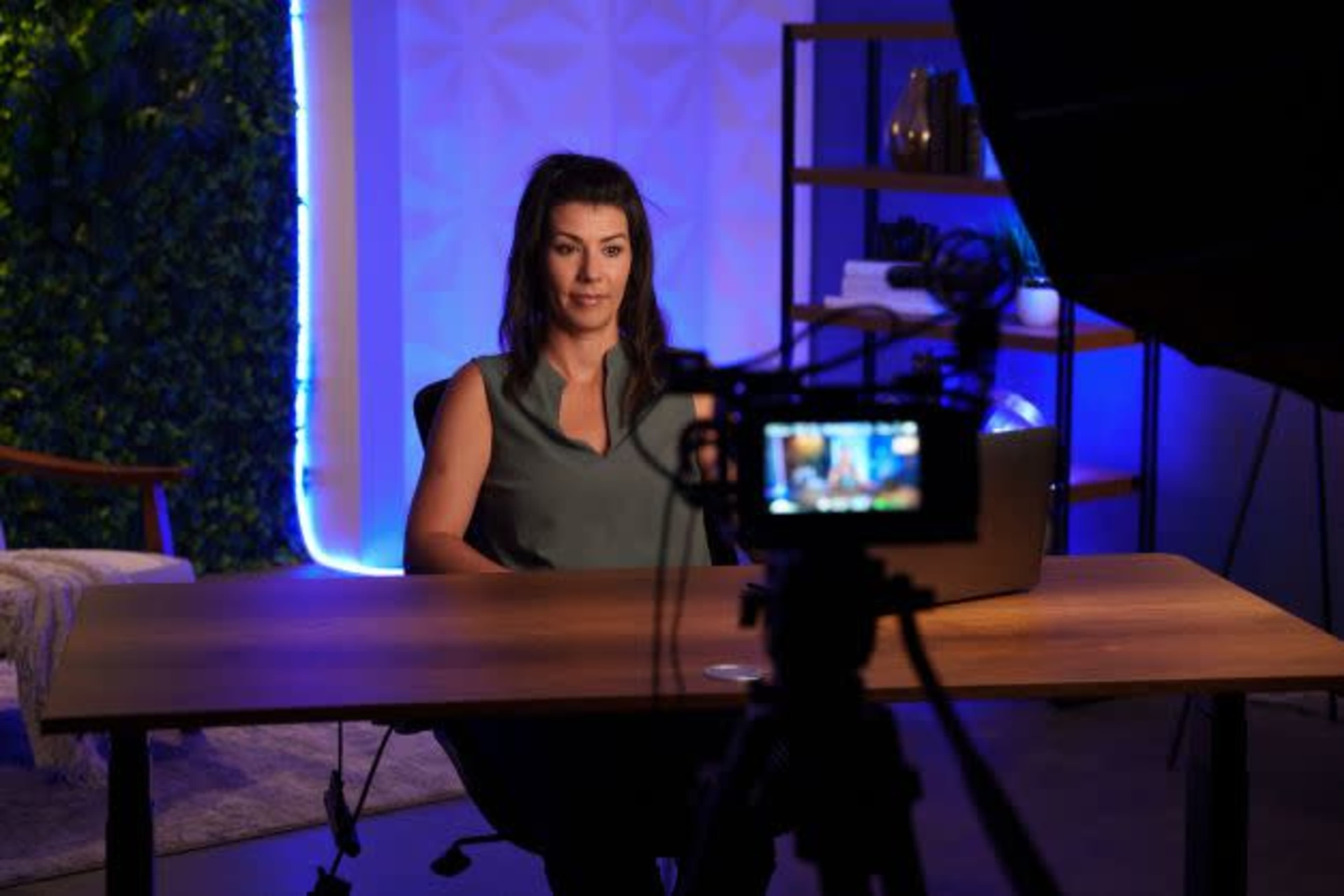 A woman sits at a desk in front of a camera, with a laptop and softly lit background in a well-decorated indoor space.
