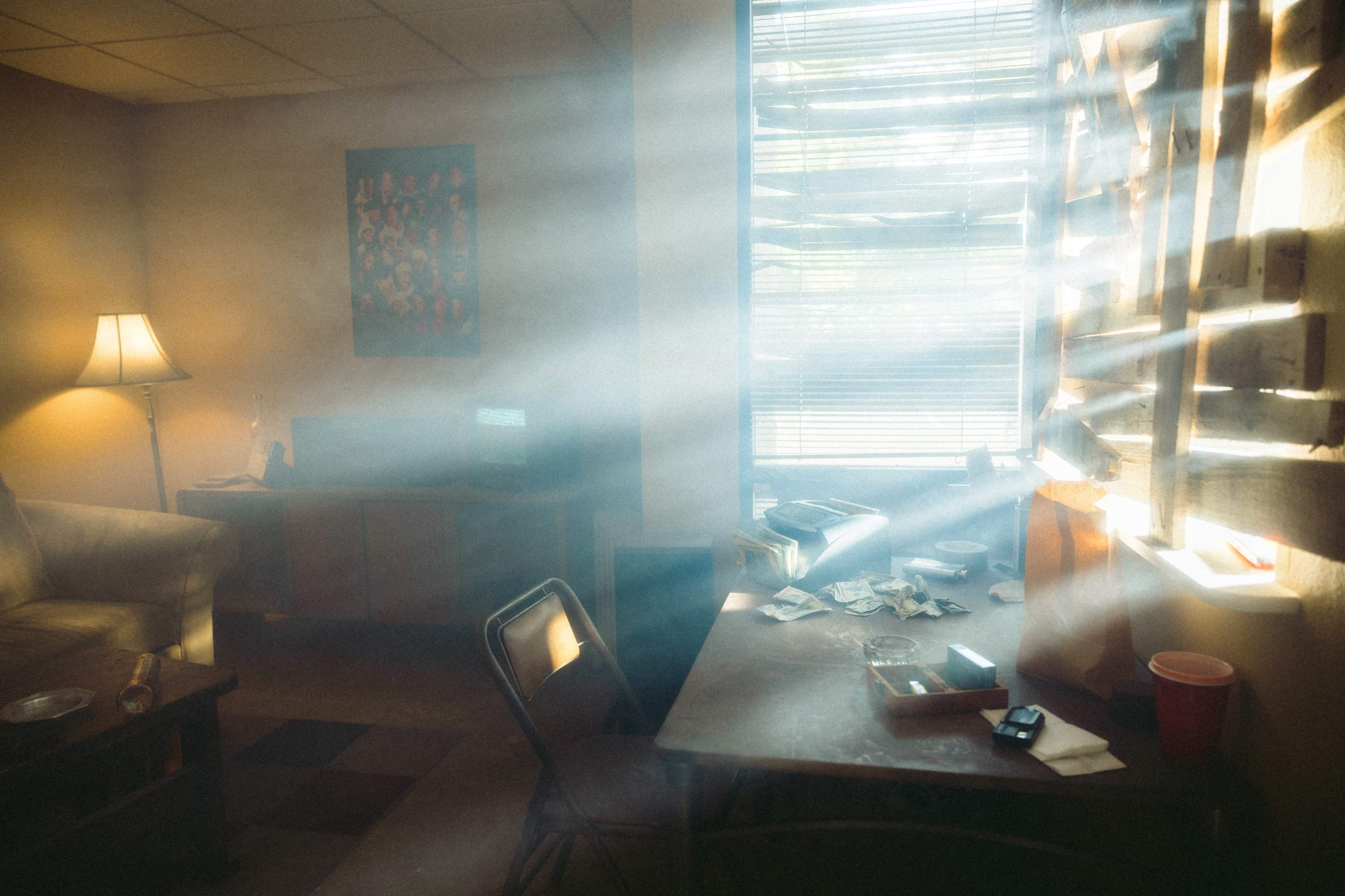 A dimly lit room with sunlight streaming through partially closed blinds, illuminating dust particles in the air and revealing a cluttered table and a couch.