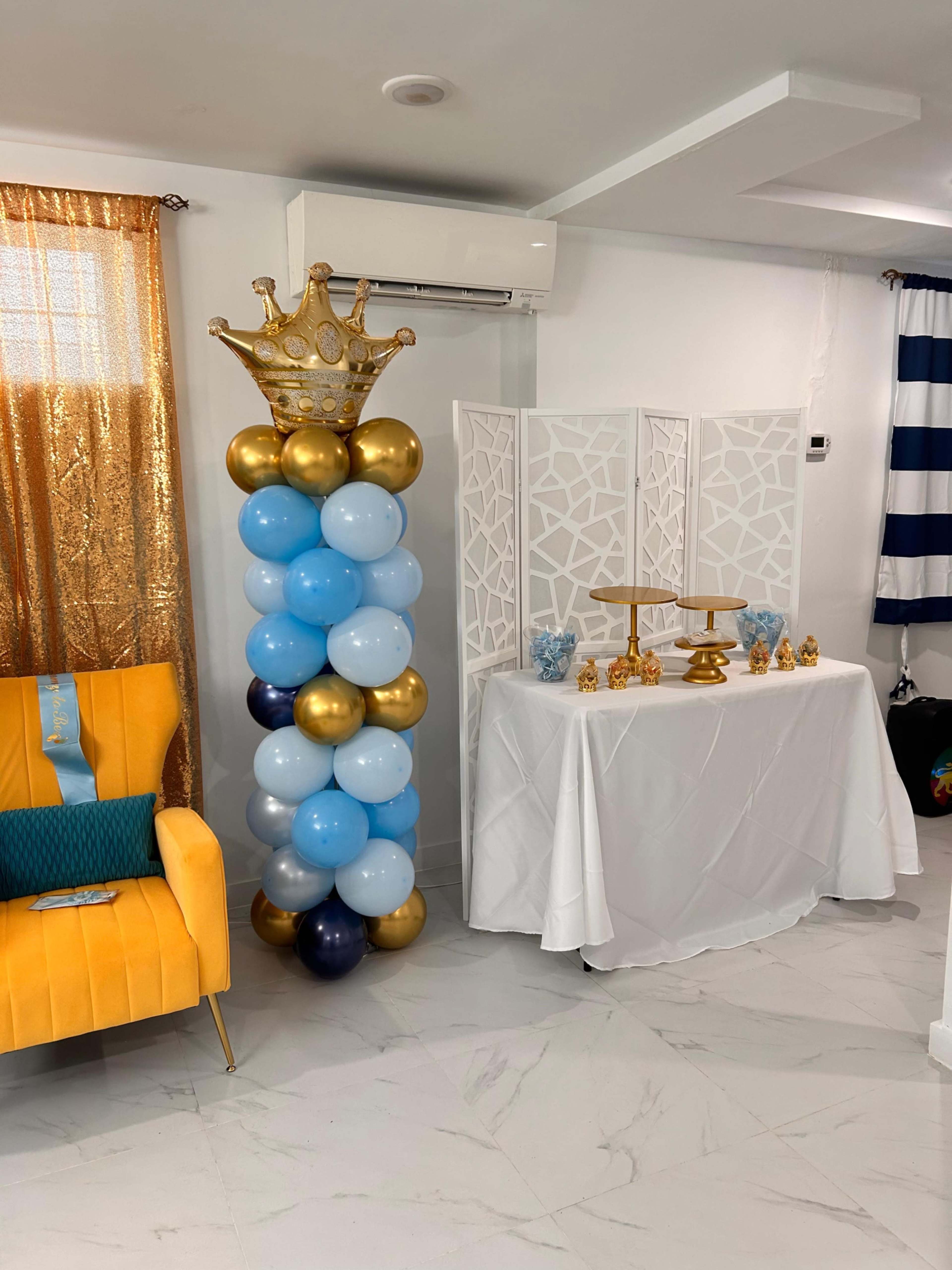 The image shows a decorated indoor space featuring a tall balloon pillar in blue, gold, and white, alongside a white table set with gold decorations and a partition screen.