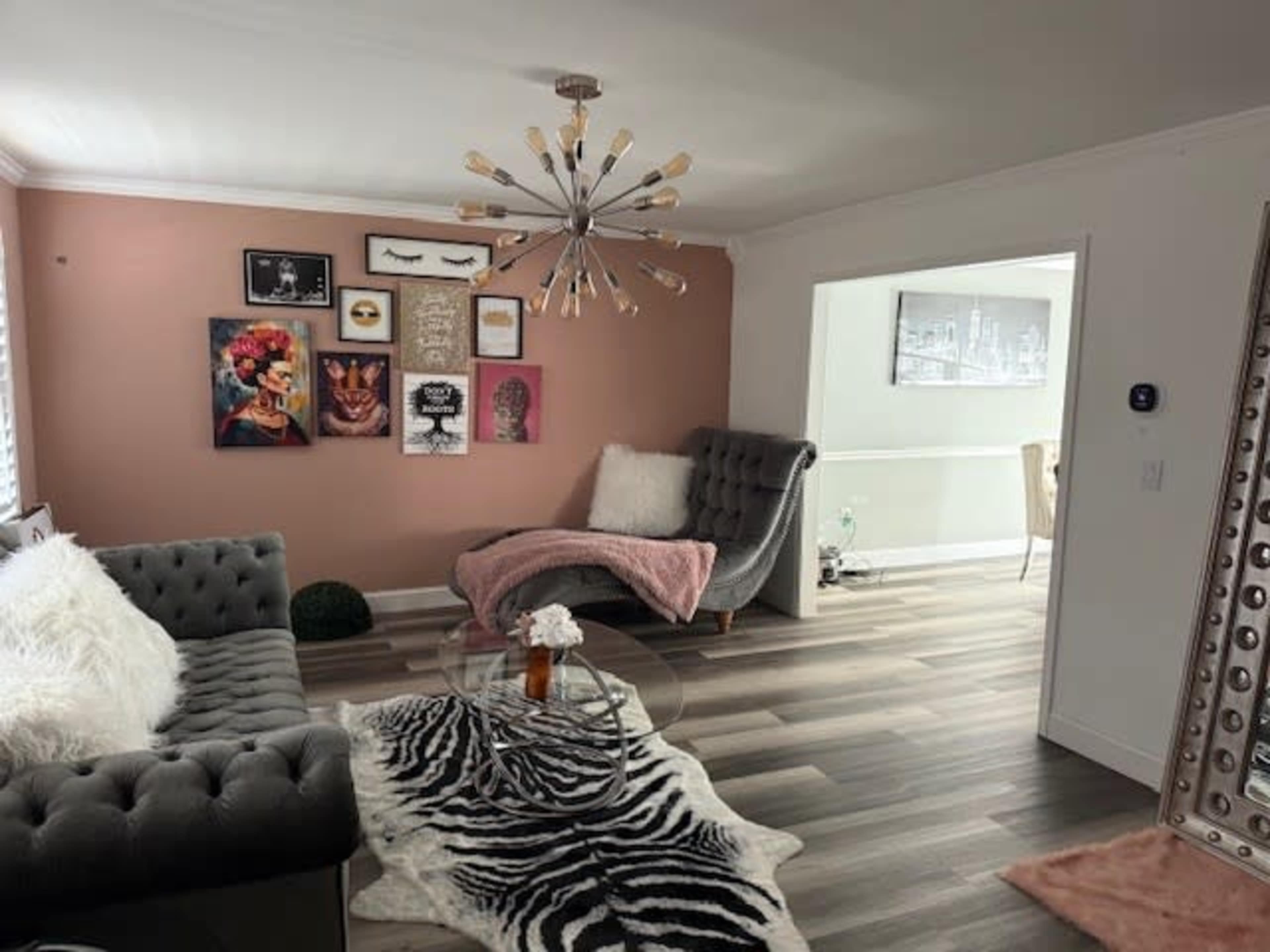 The image shows a modern living room with a plush gray sofa, a round glass coffee table, and a wall adorned with framed artwork, including a portrait.