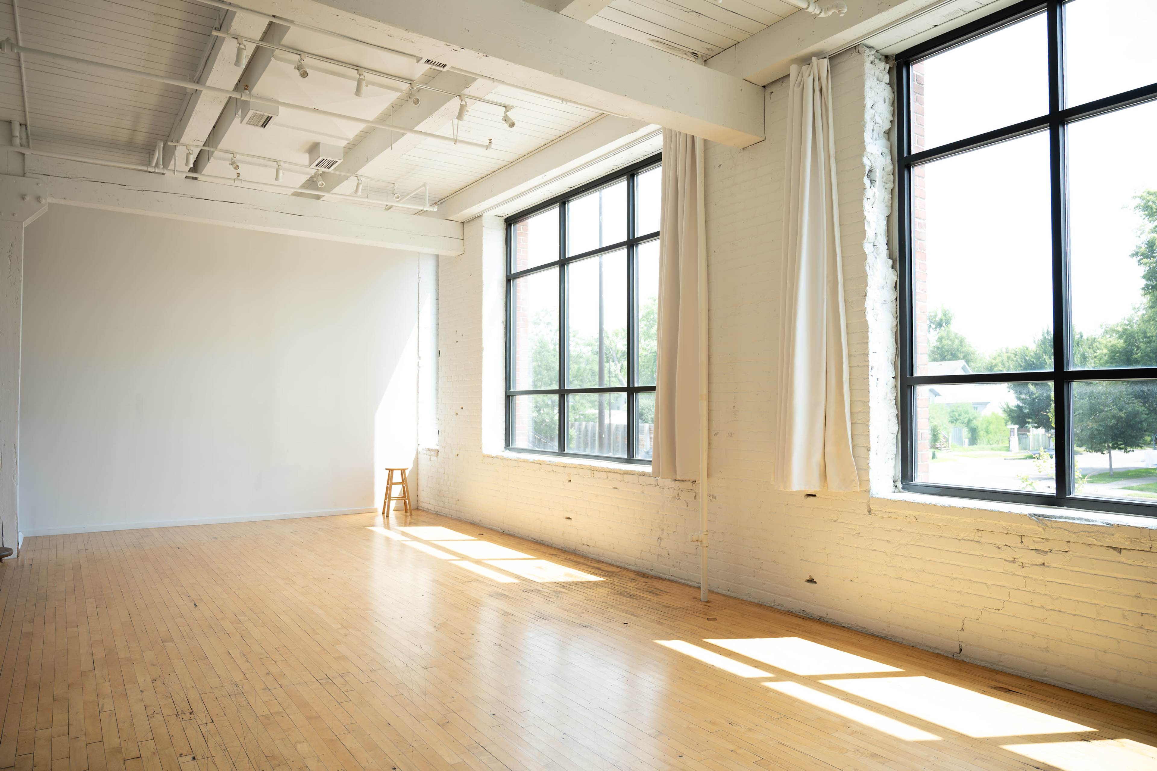 The image shows a bright, spacious room with large windows, light wooden flooring, and a minimalist aesthetic.