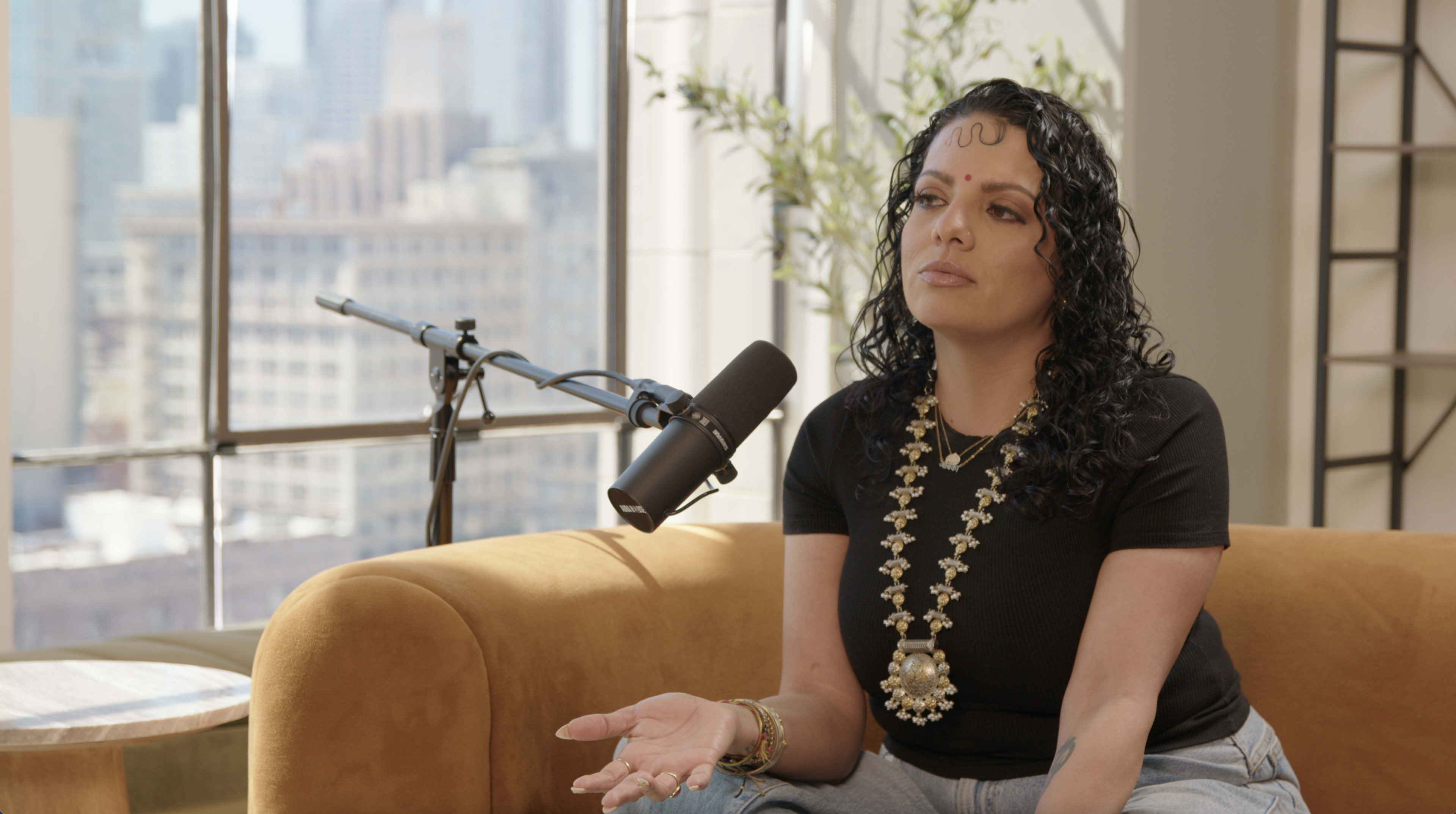 A woman with curly black hair is sitting on a tan couch in front of a microphone and a large window displaying a city skyline.