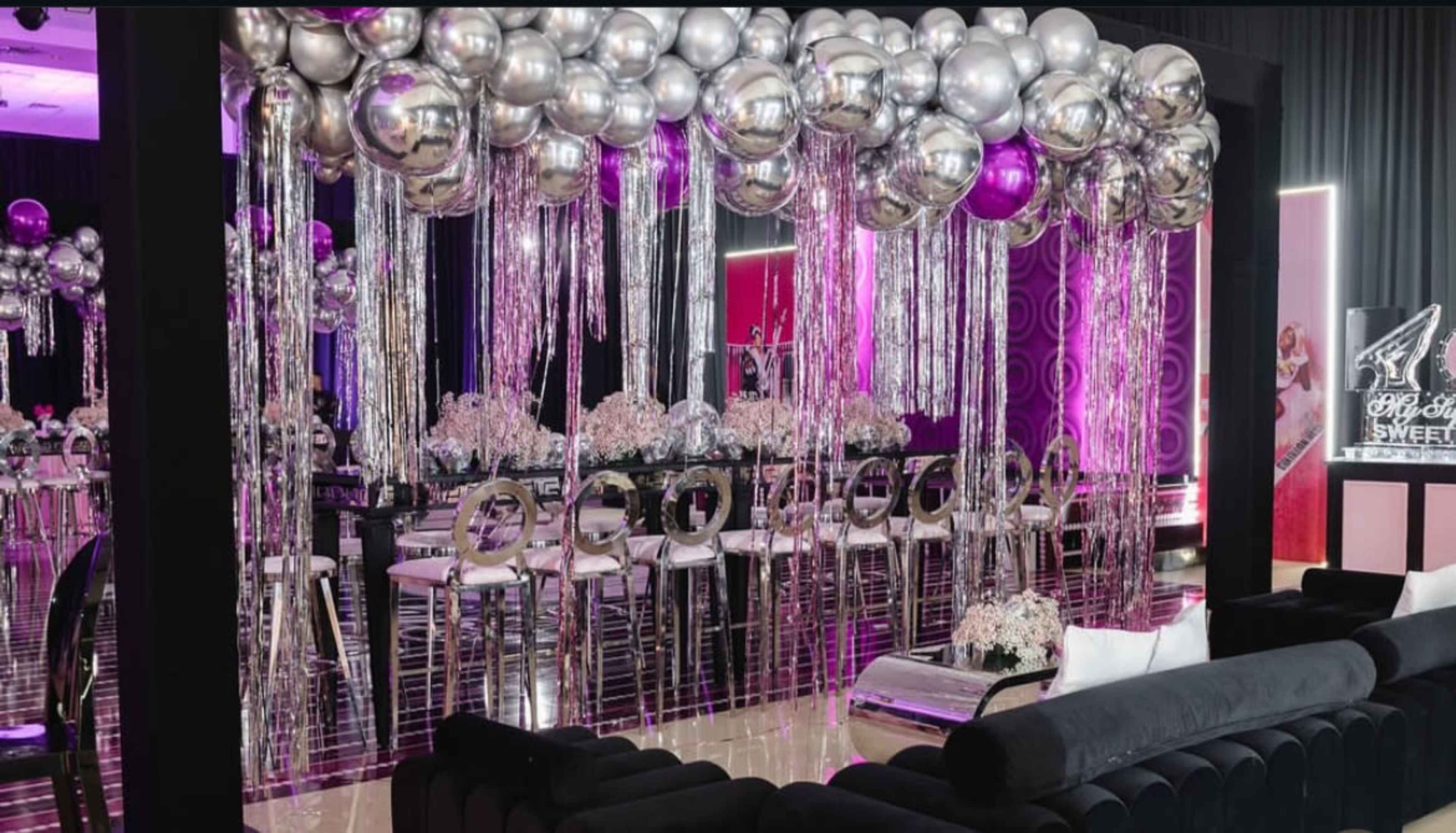 The scene features a decorated event space with metallic balloons and streamers overhead, along with elegant dining furnishings arranged for a celebration.