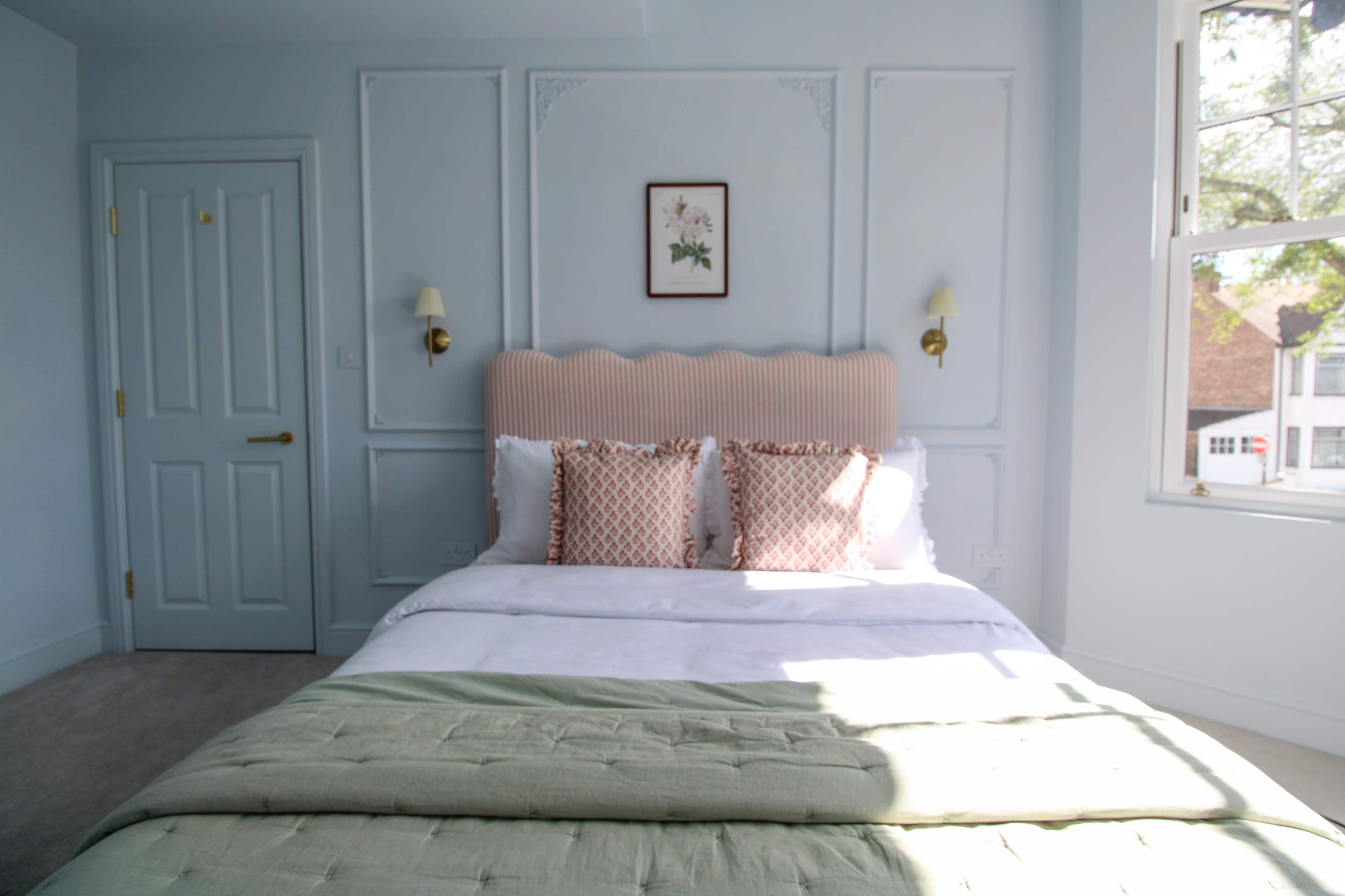 A neatly arranged bedroom features a double bed with a light green throw and decorative pillows, complemented by wall sconces and a small framed artwork.