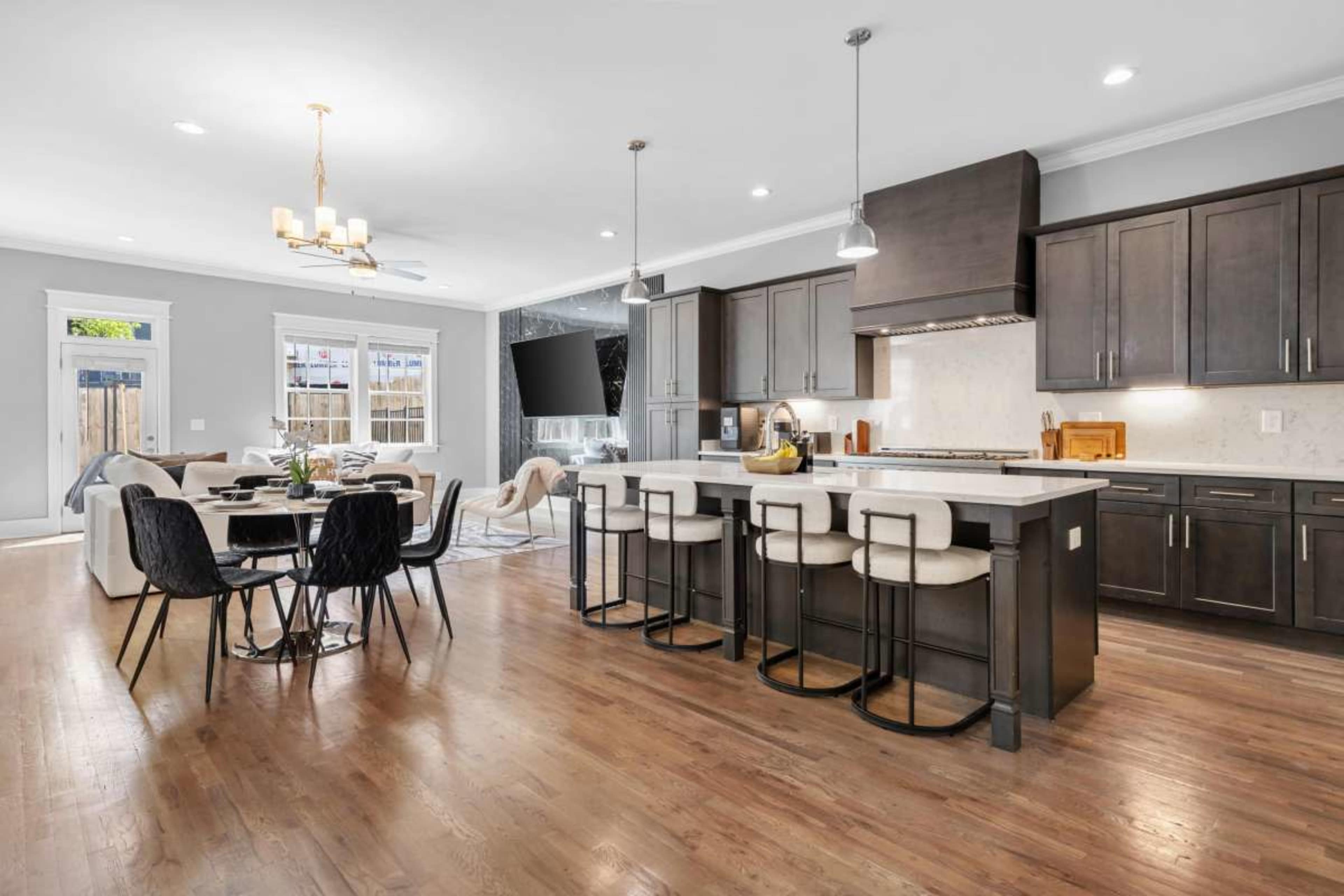 The image shows a modern open-concept kitchen and dining area featuring dark cabinetry, a large island with bar stools, and a spacious living room with a television.