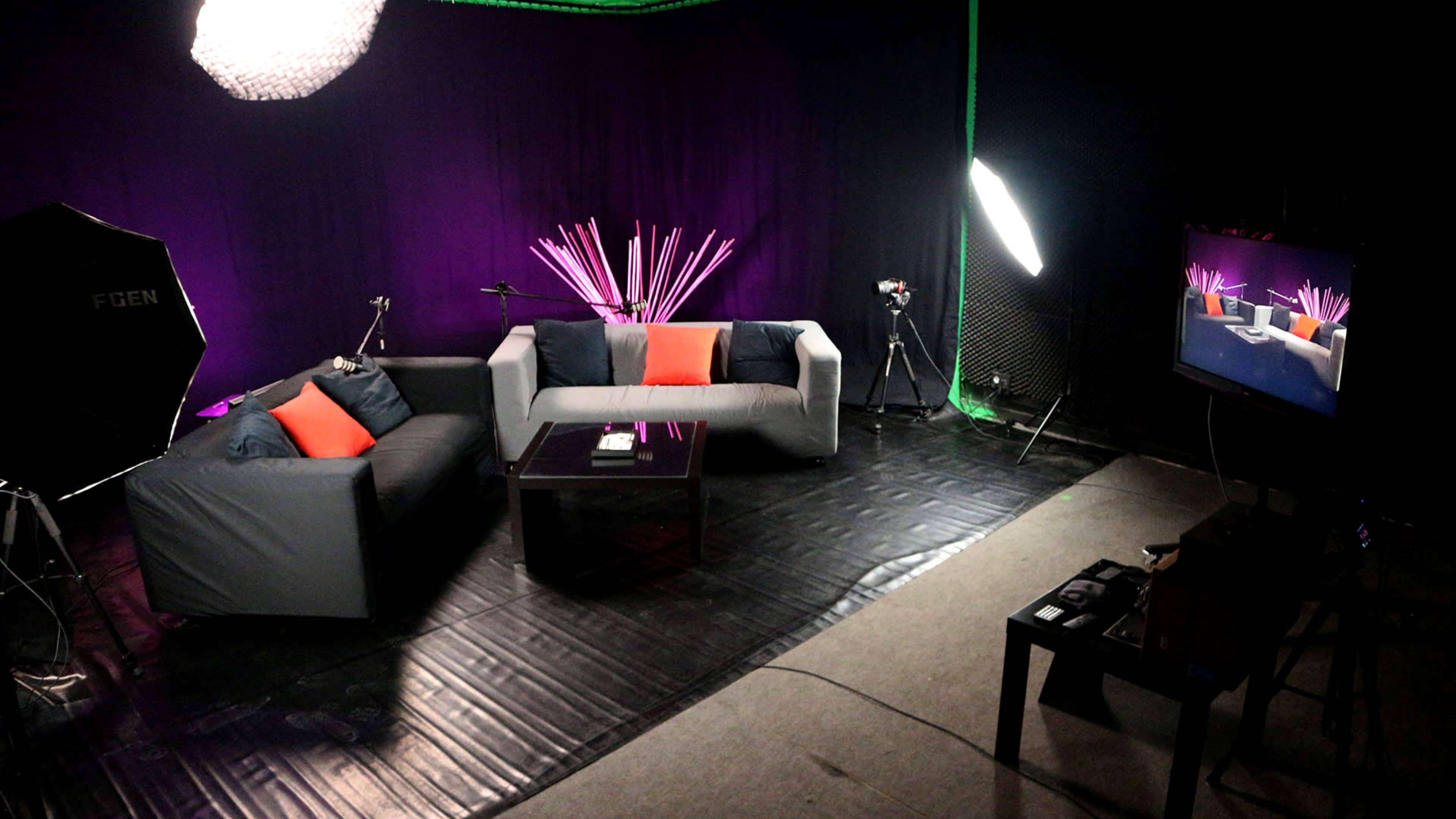 The image shows a modern studio setup with two sofas, a glass coffee table, and various lighting equipment against a purple backdrop.