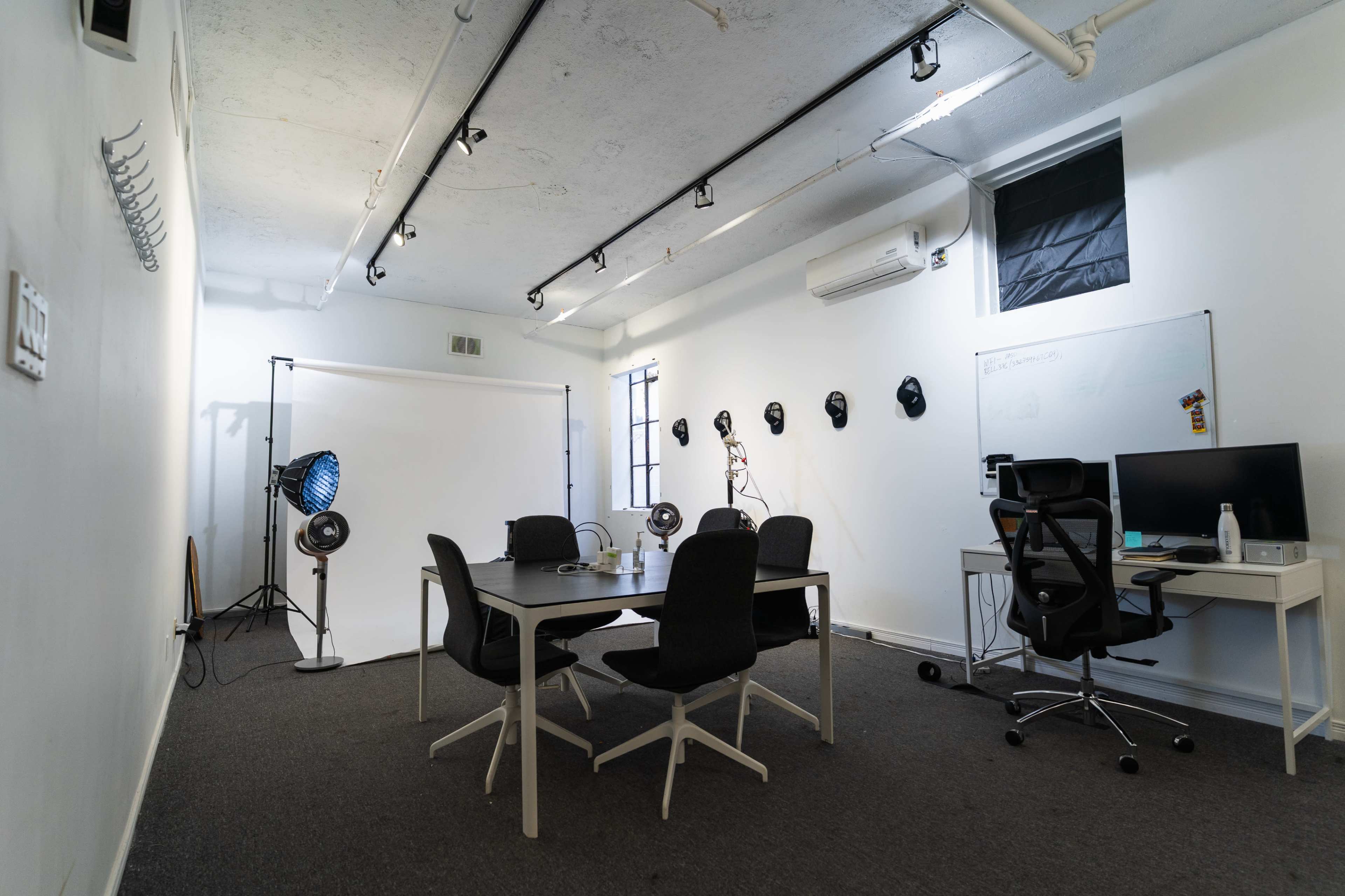 The image shows a minimalist studio space featuring a conference table, office chairs, and lighting equipment against white walls.