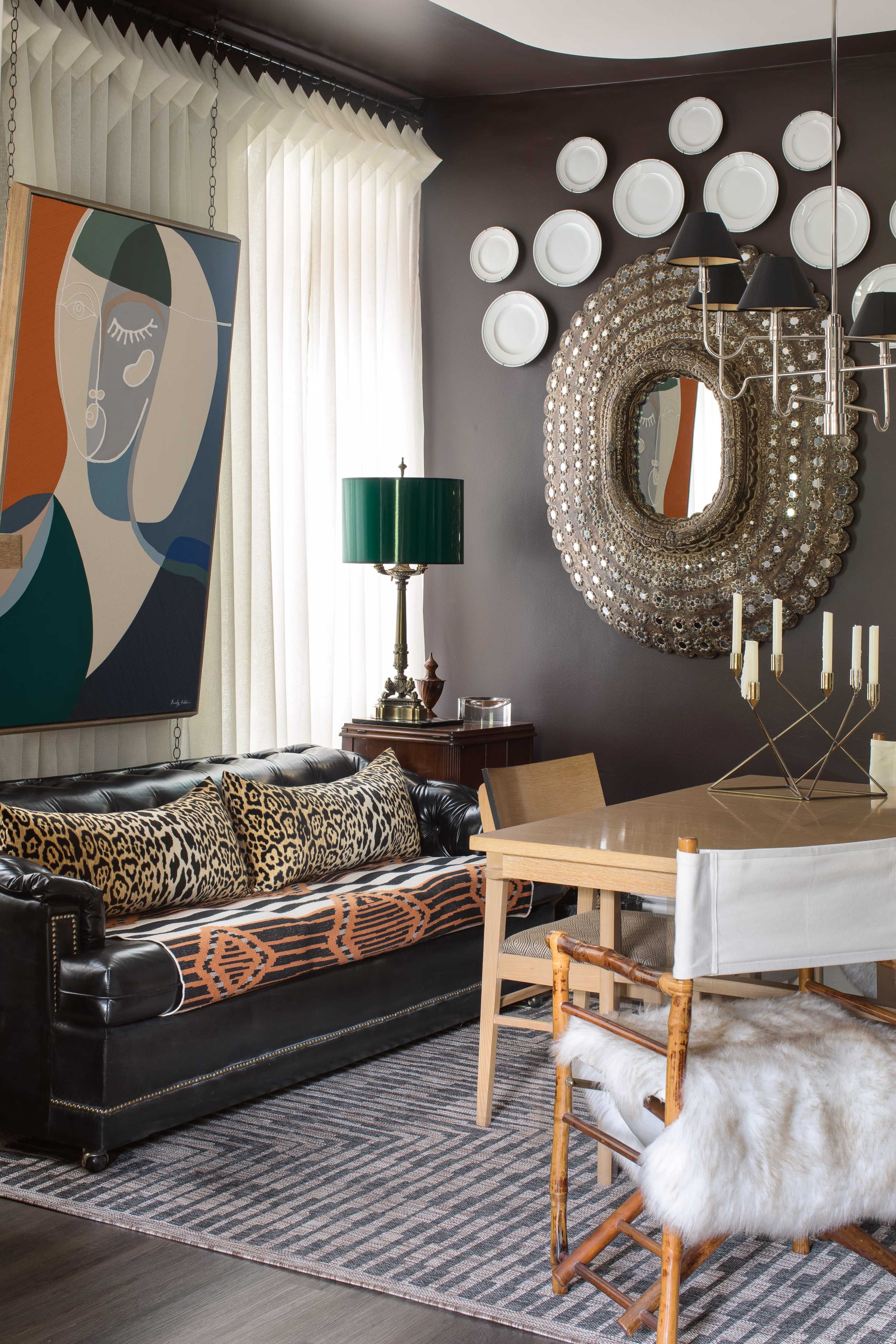 The image depicts a stylish dining area featuring a black leather sofa adorned with animal print pillows, a wooden dining table surrounded by chairs, and decorative plates displayed on a dark wall.