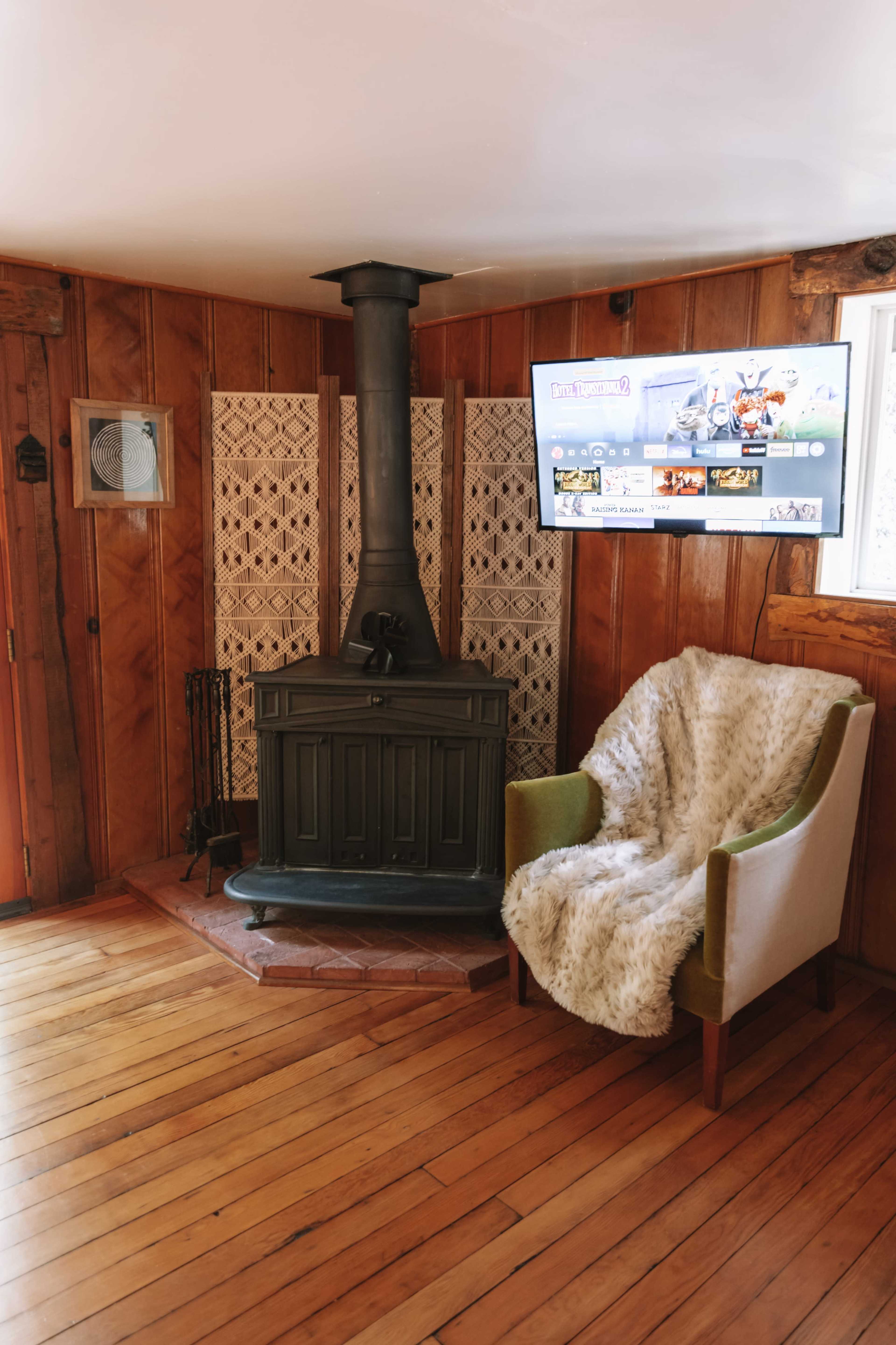 The image shows a cozy corner of a wooden room featuring a black wood stove, a green and white upholstered chair draped with a furry blanket, and a wall-mounted television.