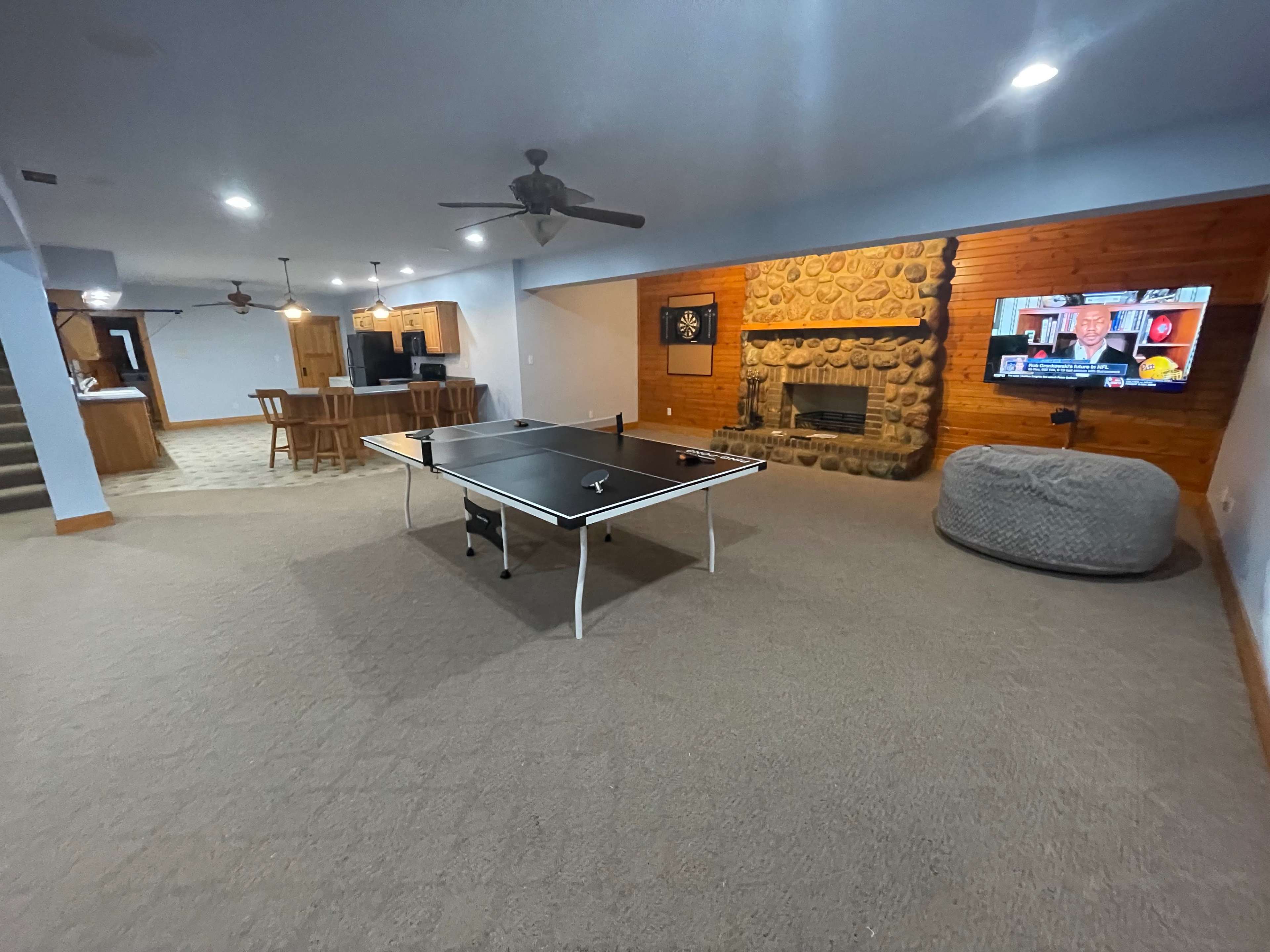 The image shows a spacious game room featuring a ping pong table, a stone fireplace, and a large screen displaying a news program.