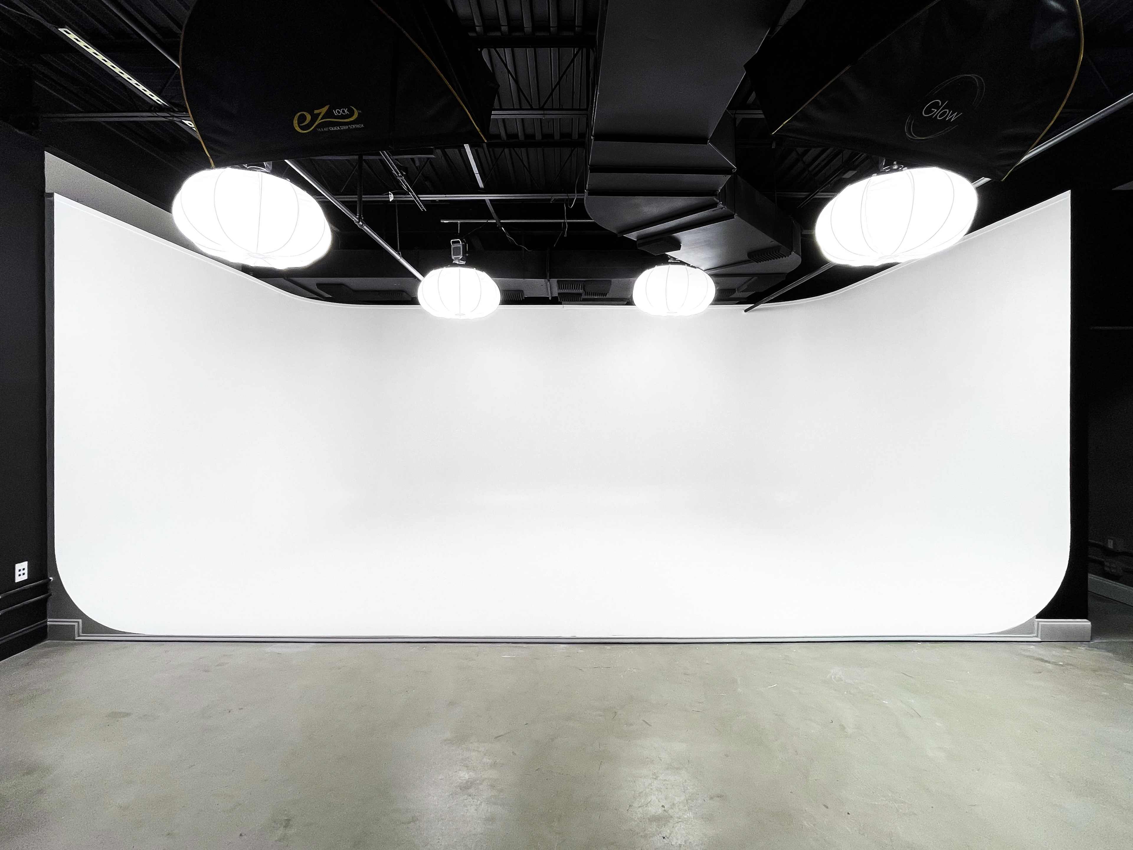 The image shows a spacious studio with a seamless white backdrop and modern lighting fixtures hanging from the ceiling.
