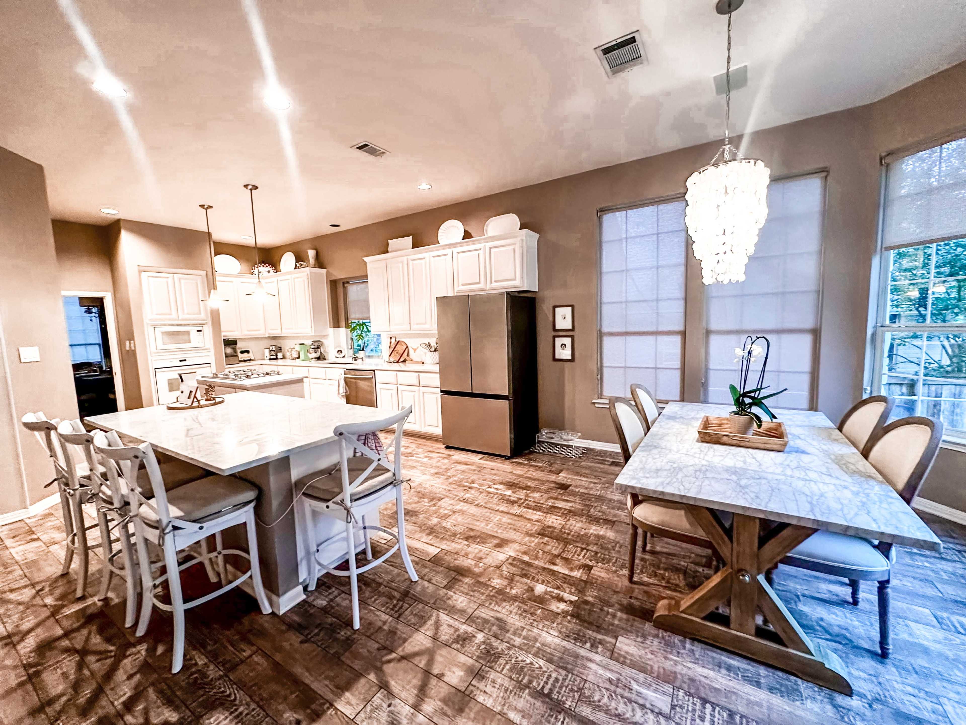 The image shows a spacious kitchen and dining area featuring a marble countertop, modern appliances, and a wooden dining table with a decorative centerpiece.