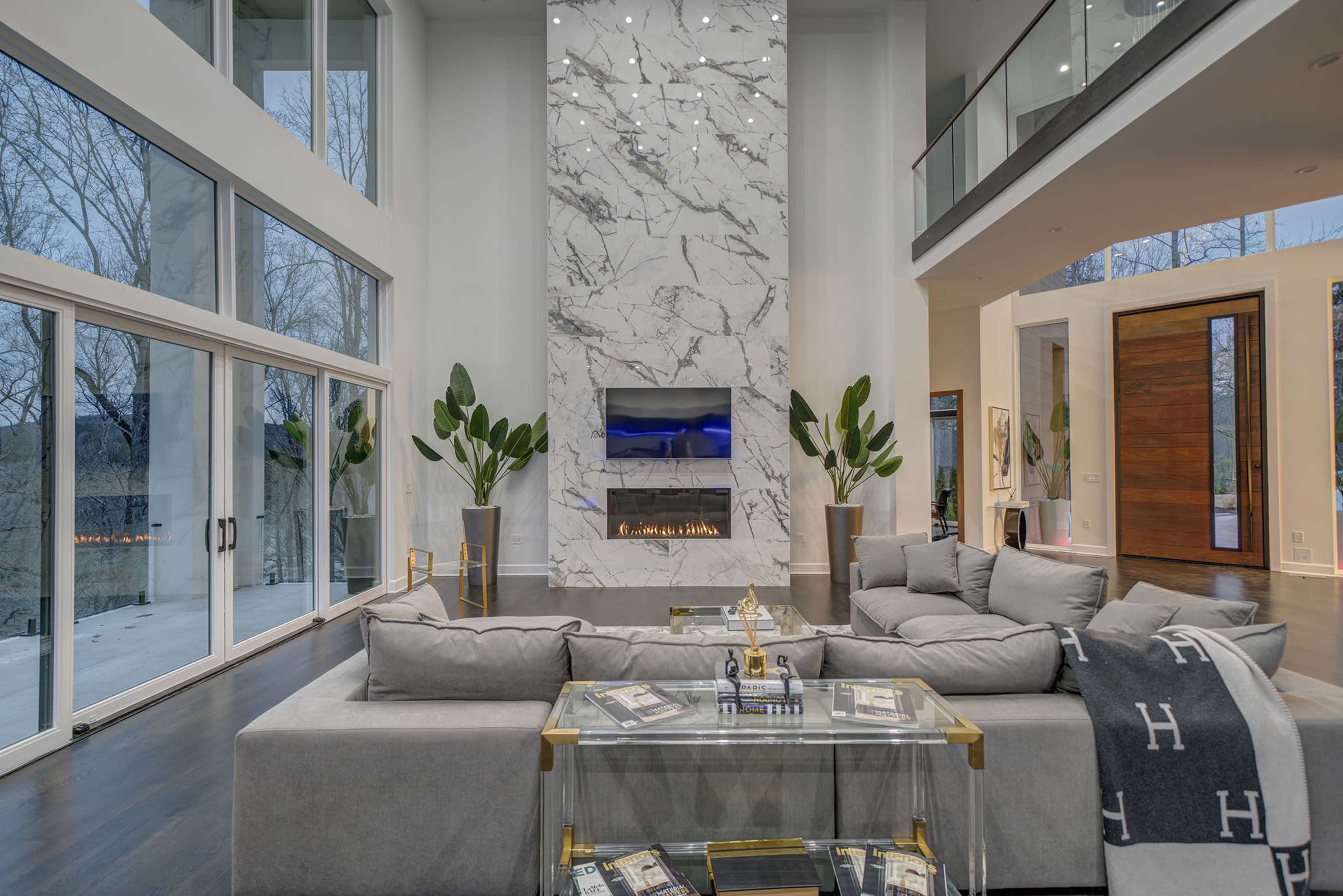 The image shows a modern living room with a large marble fireplace, floor-to-ceiling windows, and contemporary furniture arranged around a glass coffee table.