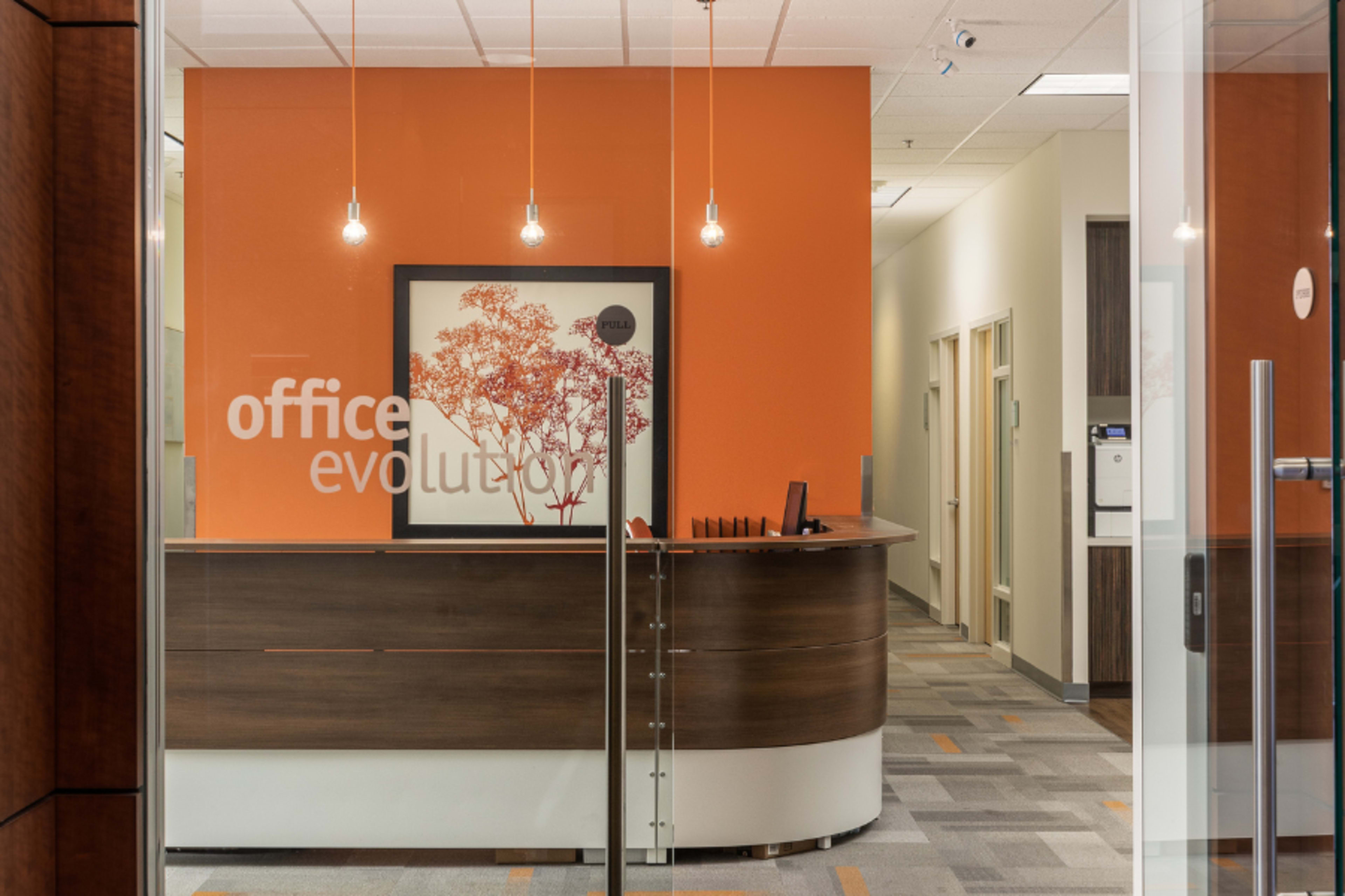 A modern office reception area with a wooden front desk, orange accent wall, and framed artwork, viewed through a glass entrance.