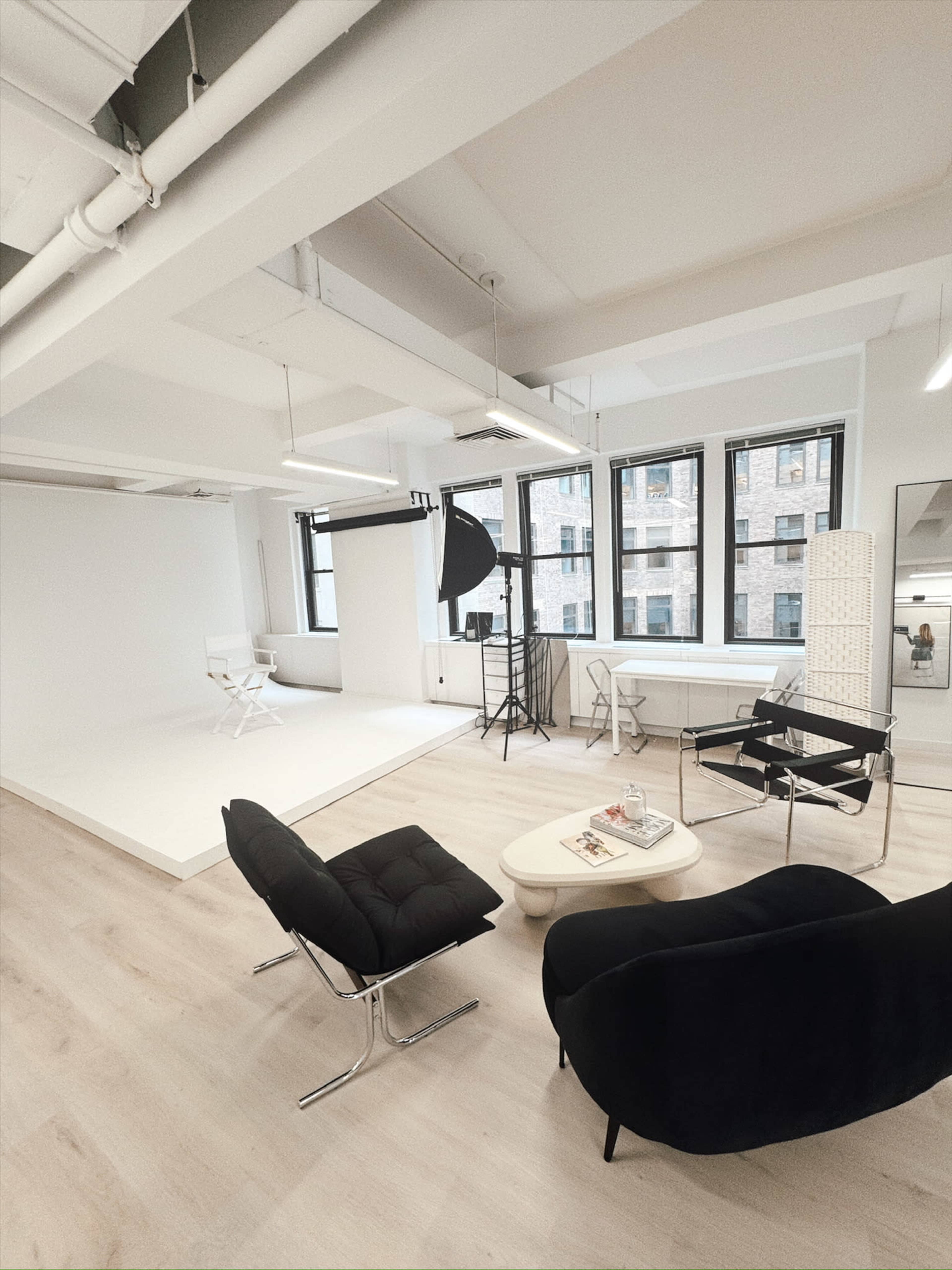 The image shows a spacious studio with modern furniture, including a black lounge chair, a white table, and a backdrop for photography.
