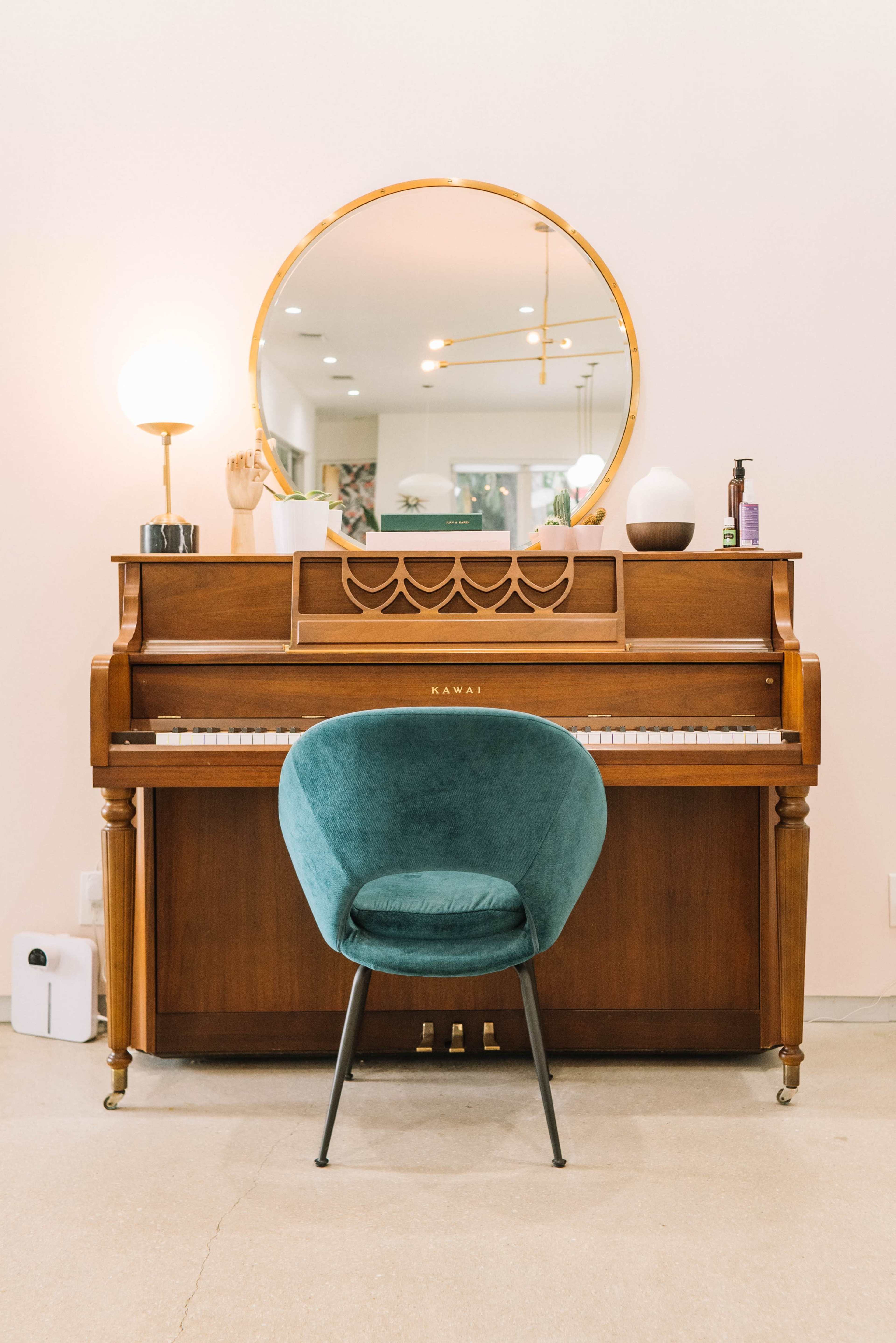 A wooden Kawai piano is positioned against a wall with a round mirror above it and a teal chair in front.