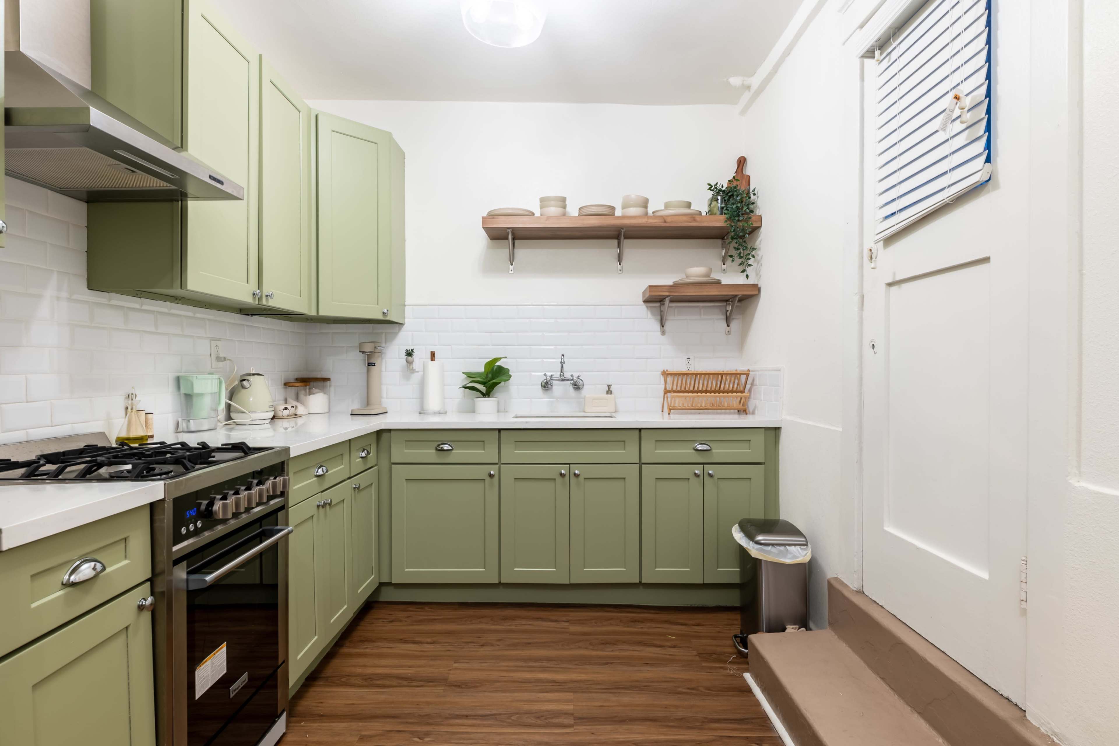 A compact kitchen features green cabinetry, a stainless steel gas stove, and wooden shelves with various kitchen items.