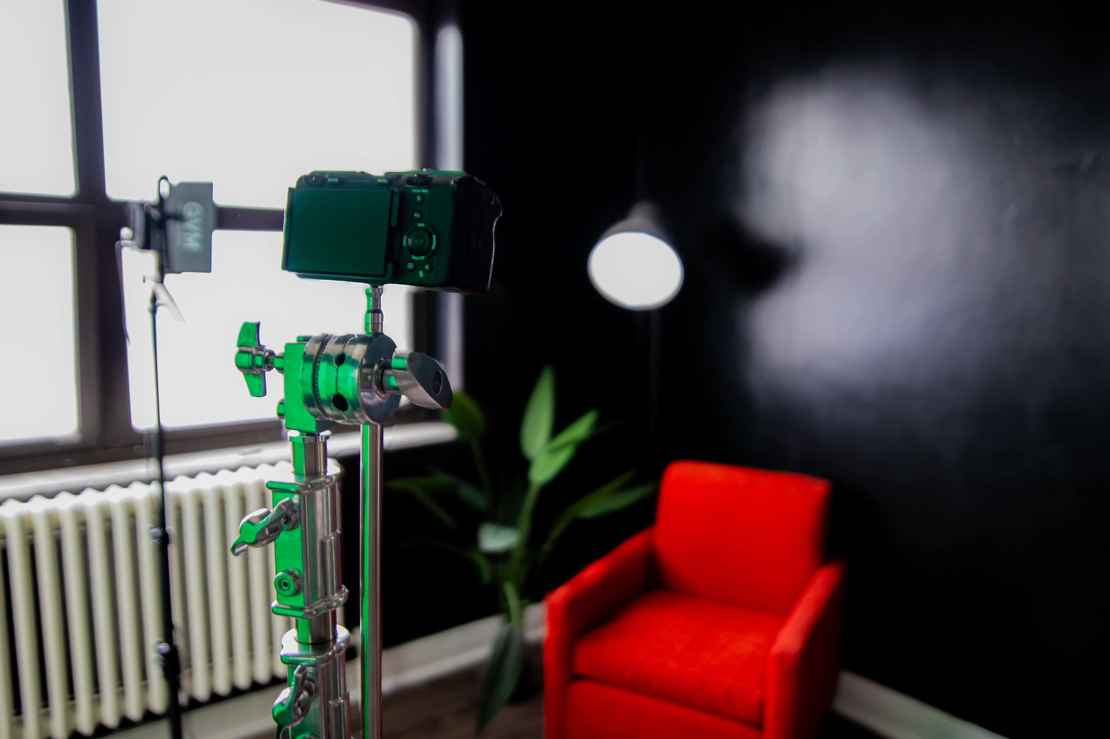 The image features a camera on a green tripod in a room with a black wall, an orange chair, a standing lamp, and green foliage.