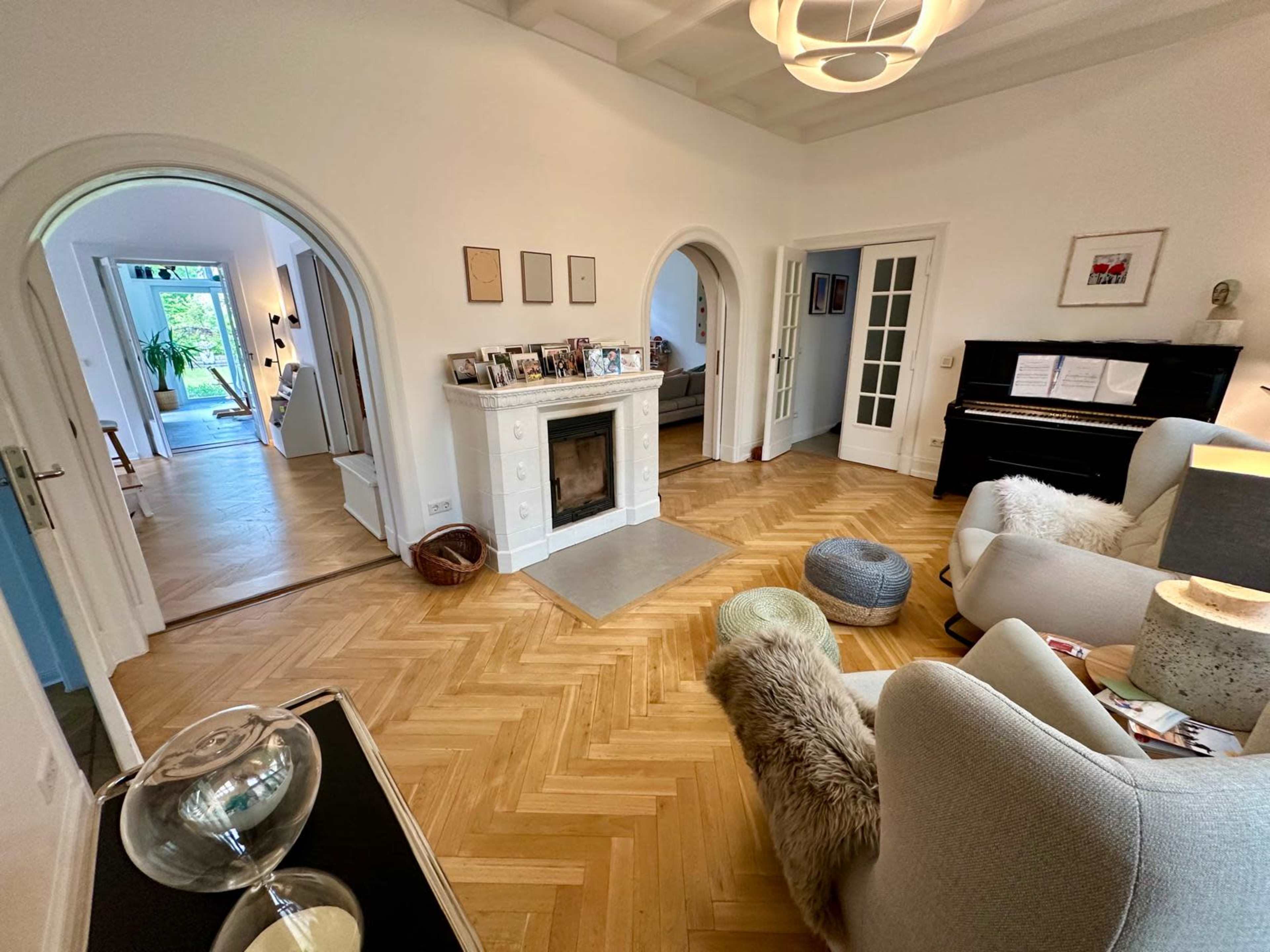 The image shows a bright living room with light-colored furniture, a piano, and a herringbone-patterned wooden floor, leading to arched doorways that connect to other areas of the house.