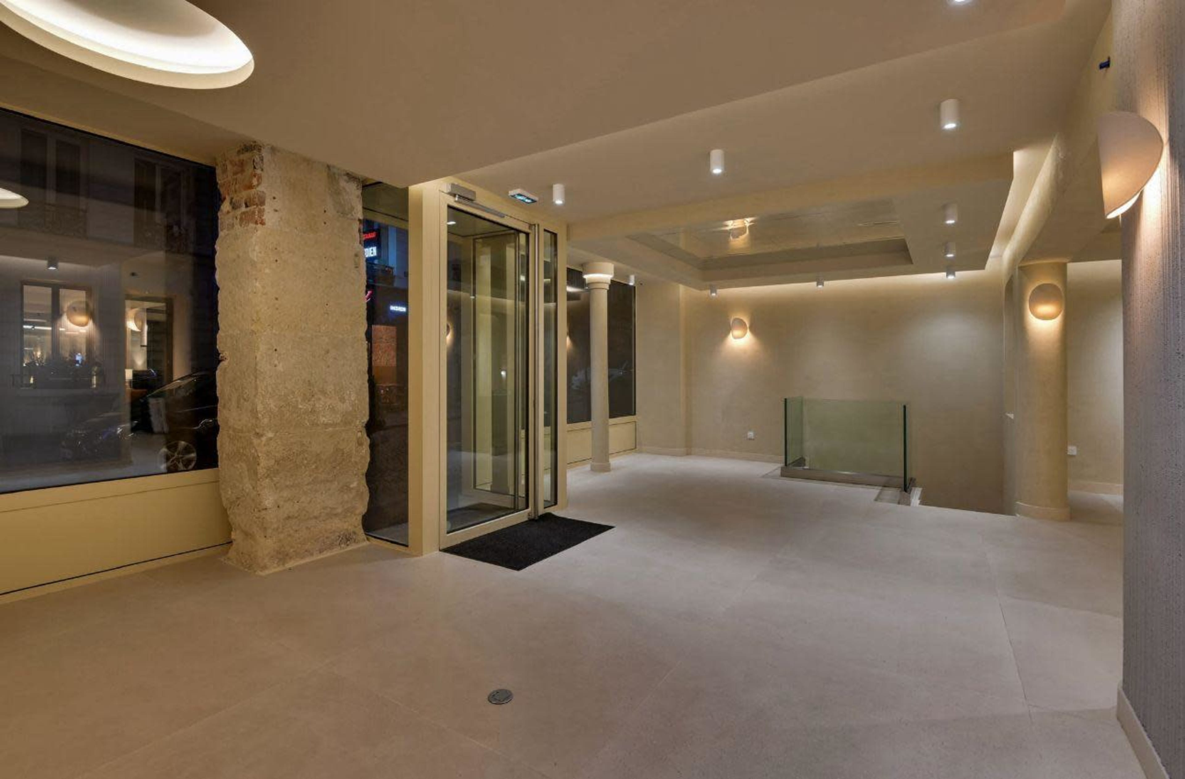 The image shows a modern, empty lobby with large glass doors, beige walls, and recessed lighting.