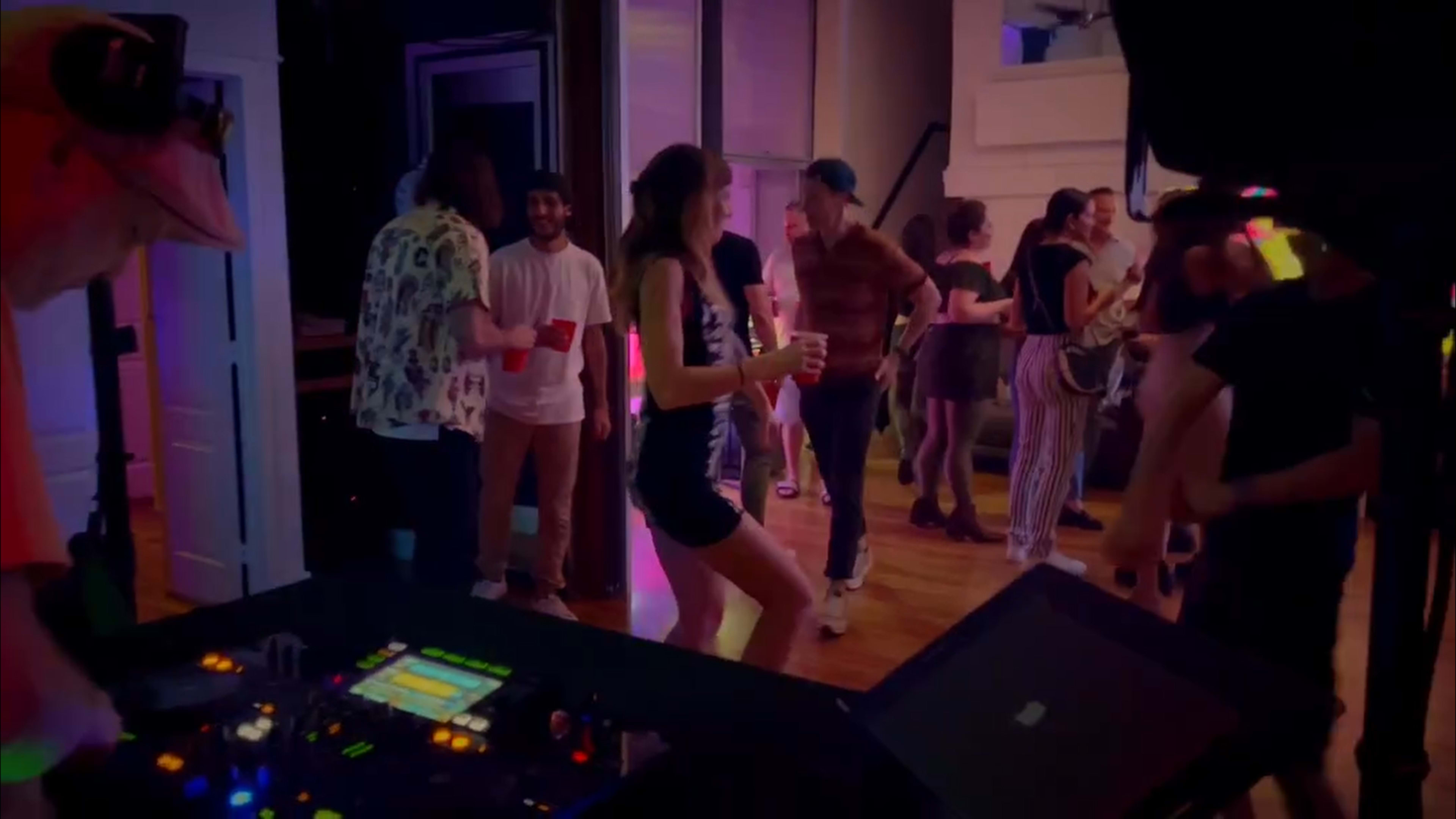 A group of people dance and socialize at a party, with a DJ setup visible in the foreground.