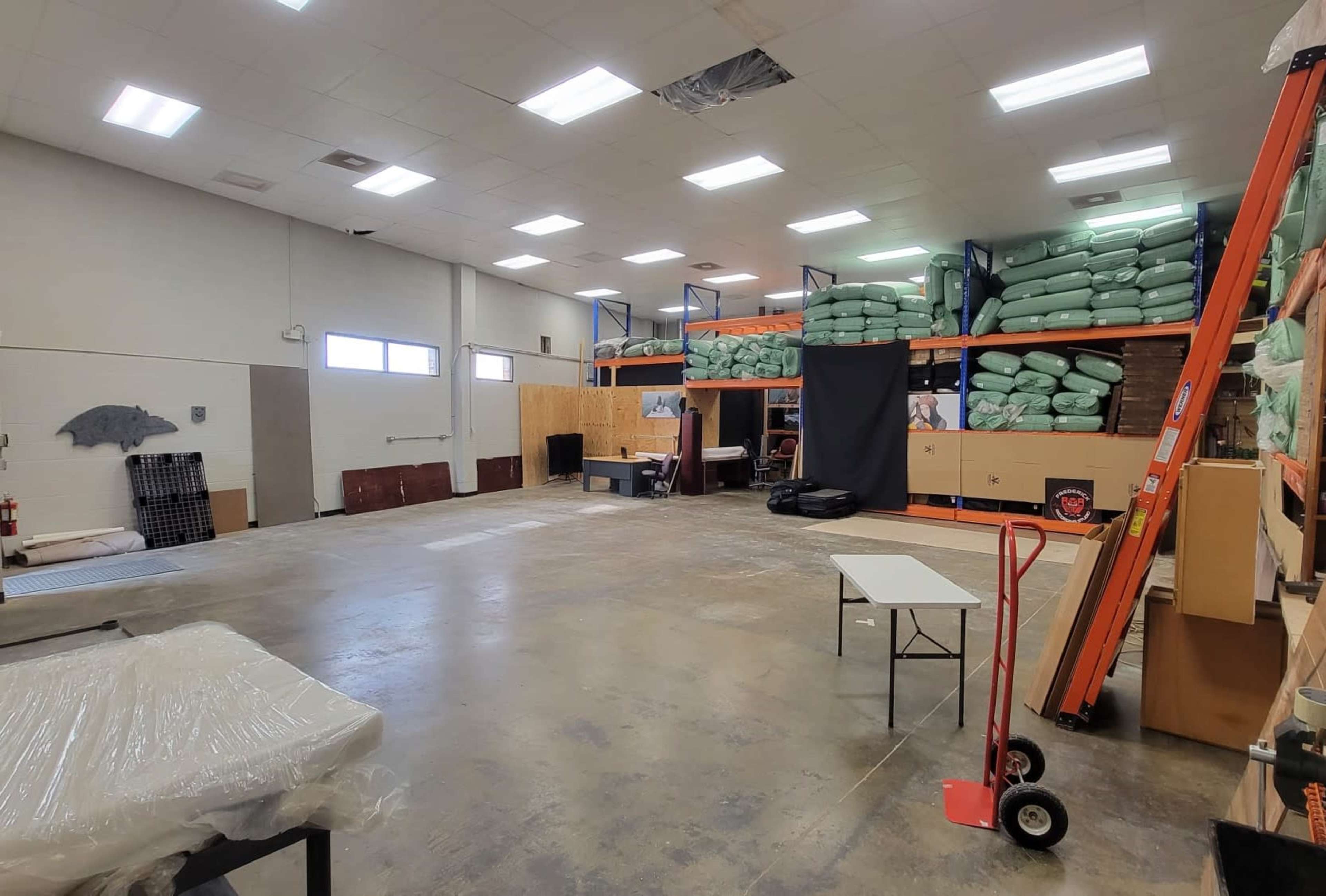 A spacious warehouse interior with shelves filled with green bags, a black curtain, and a table next to a hand truck.