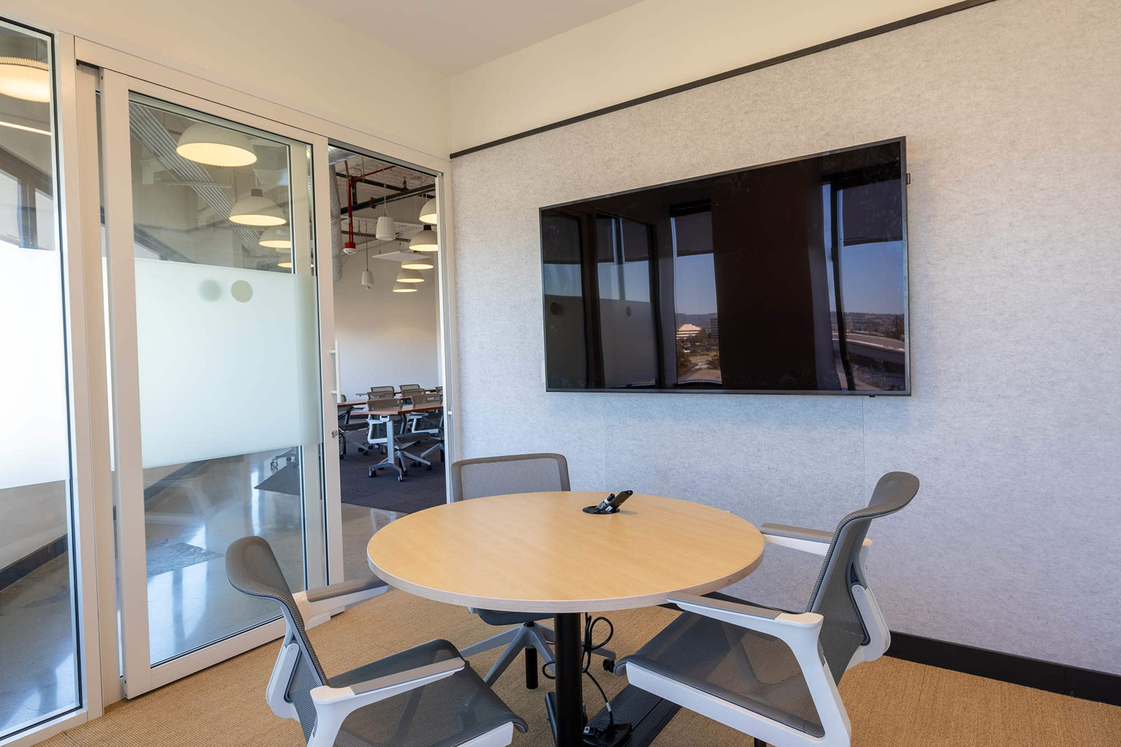 A small conference room features a round table with two chairs and a wall-mounted television, along with a glass door leading to another workspace.