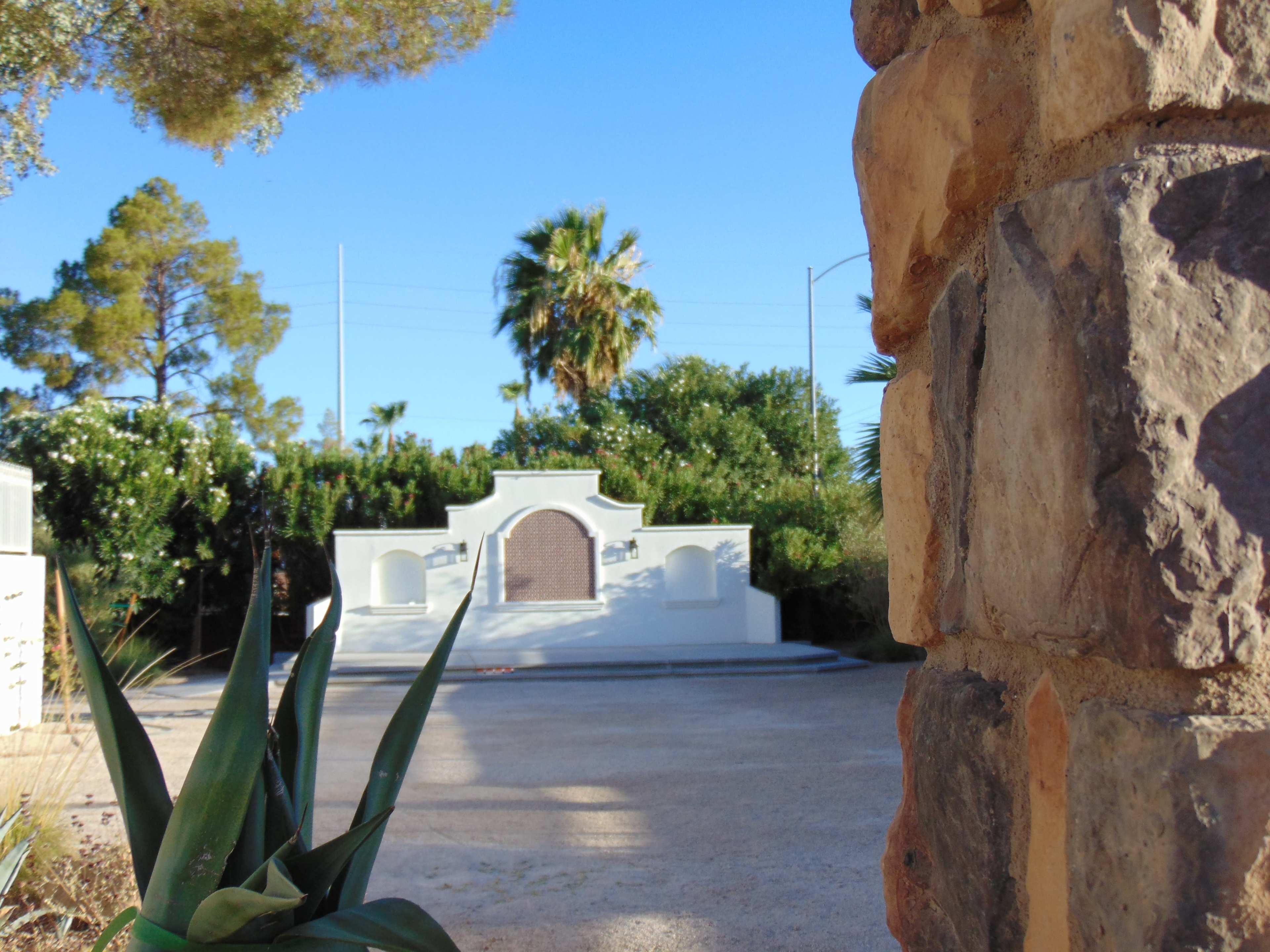 A stone wall frames a landscaped area with a white structure featuring an ornate arch and palm trees in the background.