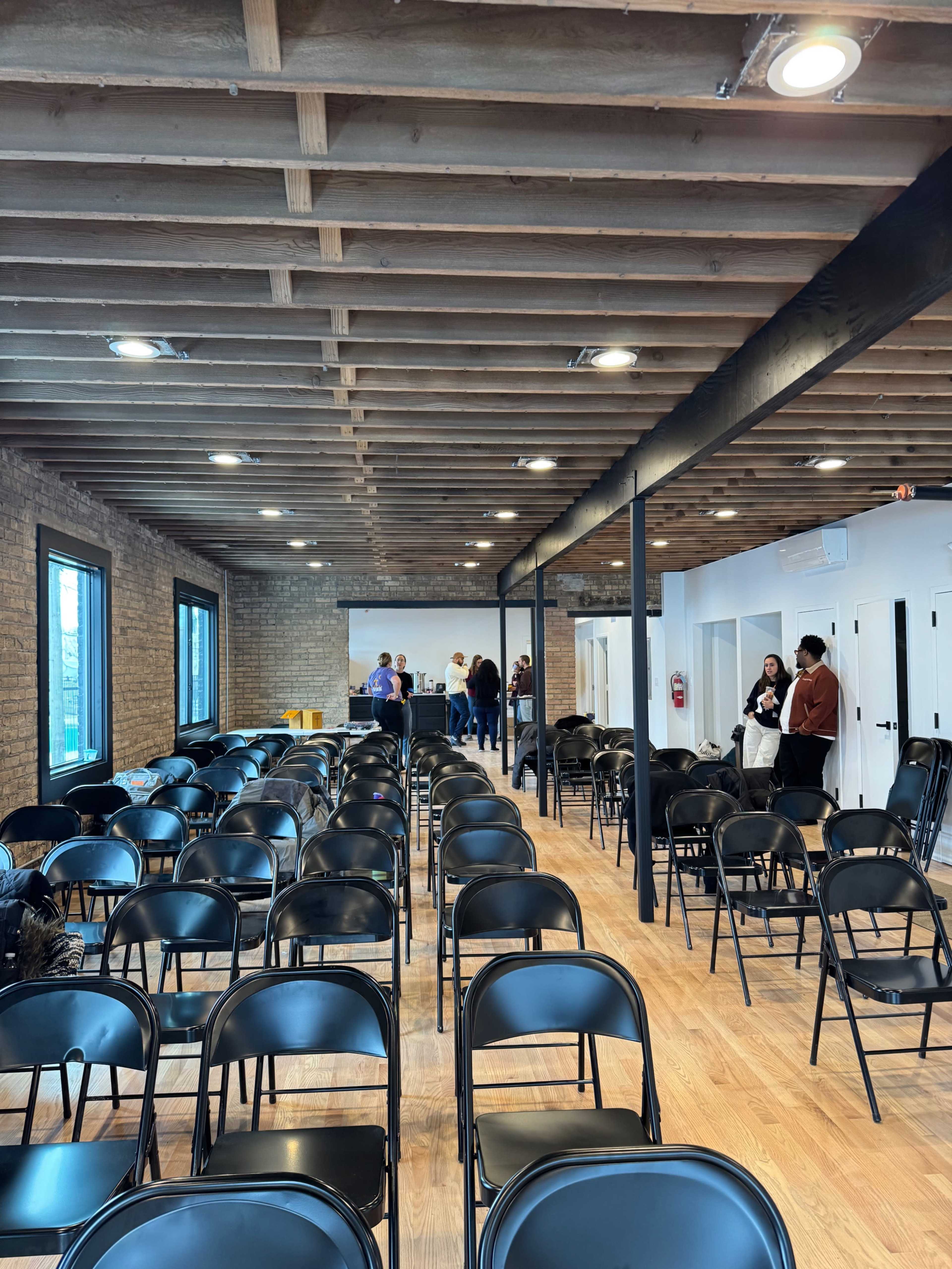 A spacious room with wooden beams and black folding chairs arranged in rows, while a small group of people converse at the far end near a screen.