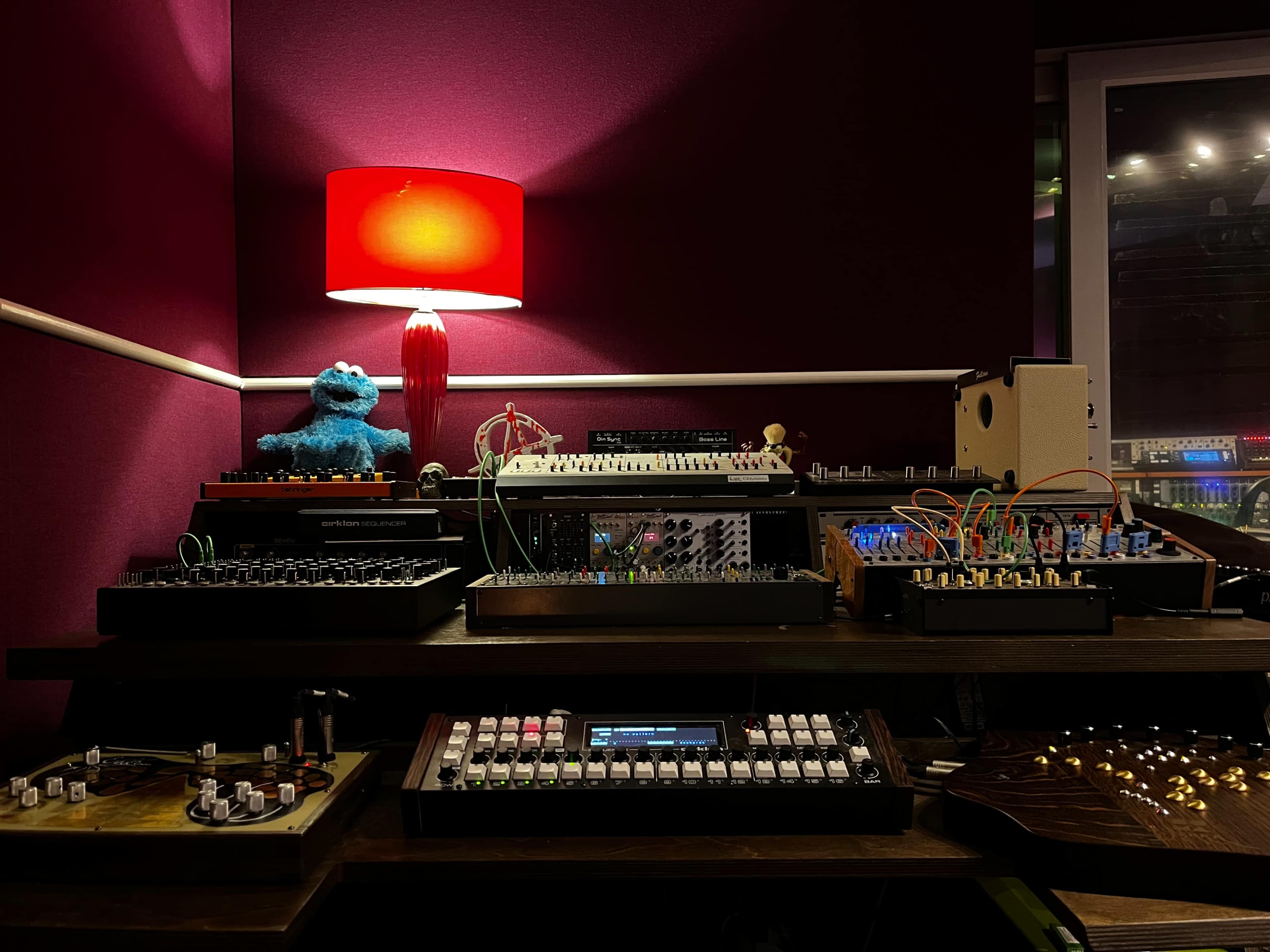 The scene features a collection of electronic musical instruments and equipment arranged on a wooden table, illuminated by a red lamp.