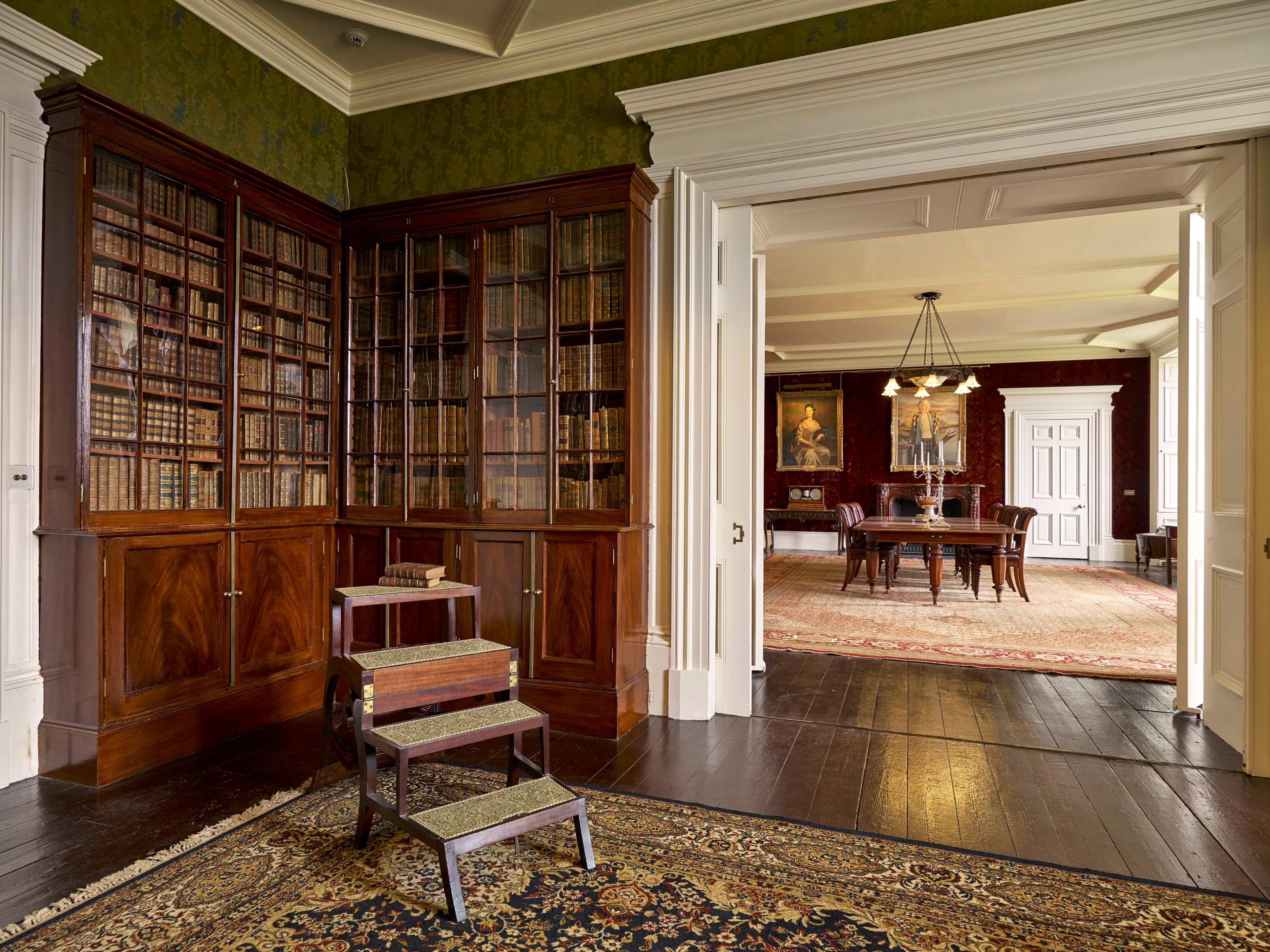 The image shows a spacious library area with wooden bookshelves filled with books, leading to a dining room featuring a wooden table and chandelier.
