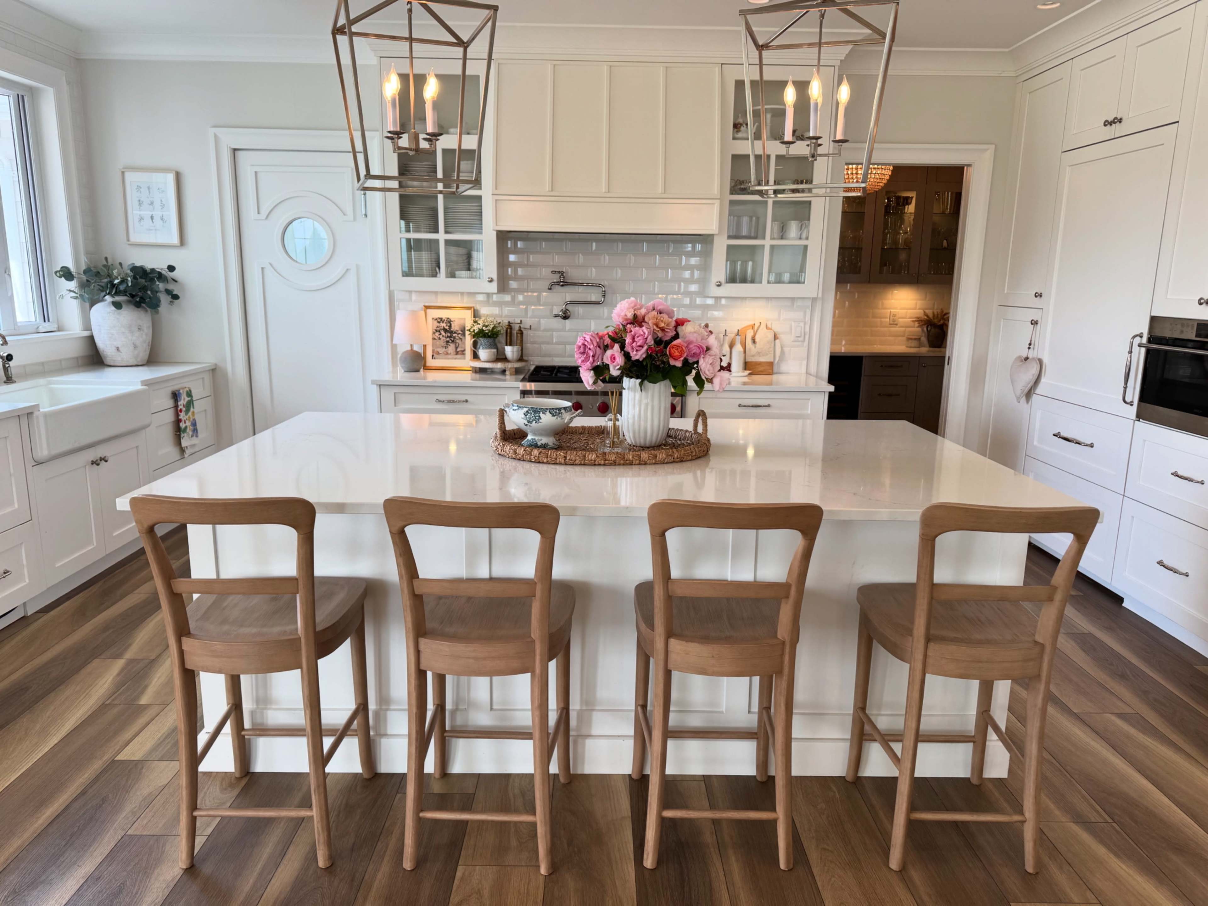 The image shows a modern kitchen with a central island featuring four wooden stools, a vase of flowers, and pendant lights hanging above.