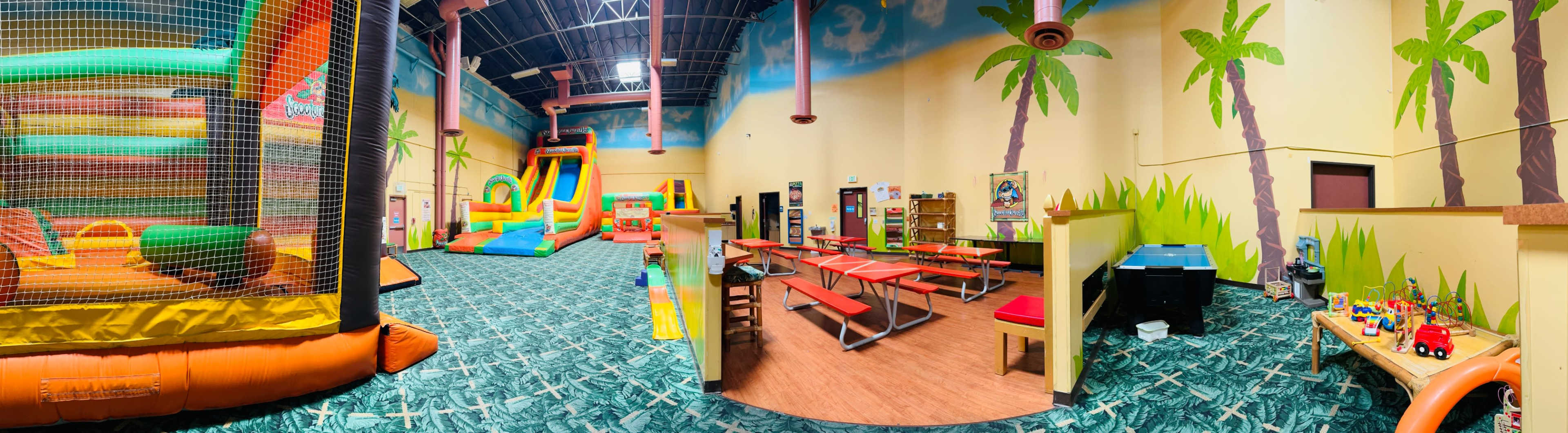 The image shows an indoor play area featuring a bounce house, slides, vibrant decorations with palm trees, tables with chairs, and various toys scattered around.
