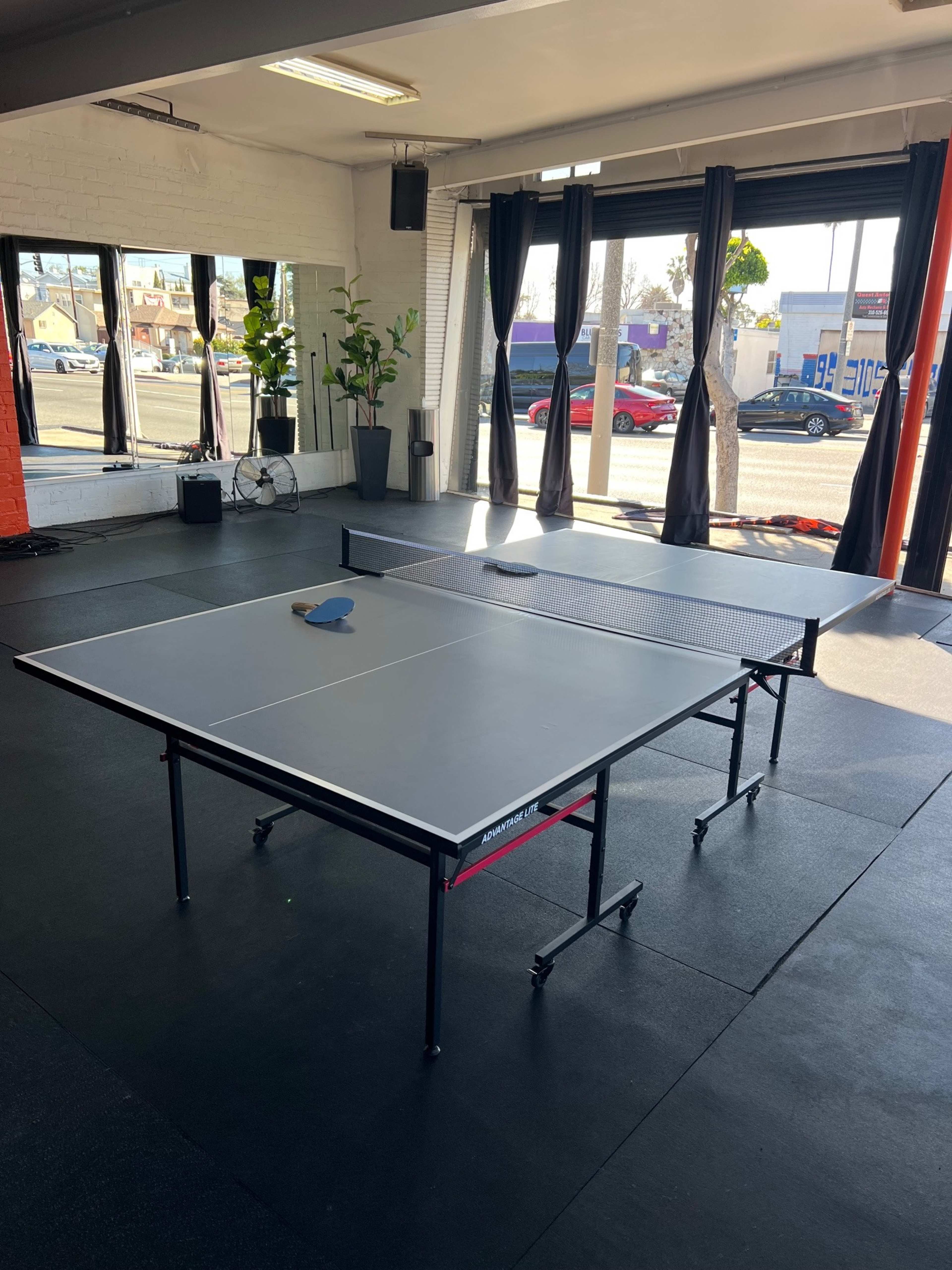 The image shows two ping pong tables set up in a brightly lit room with large windows and black curtains.