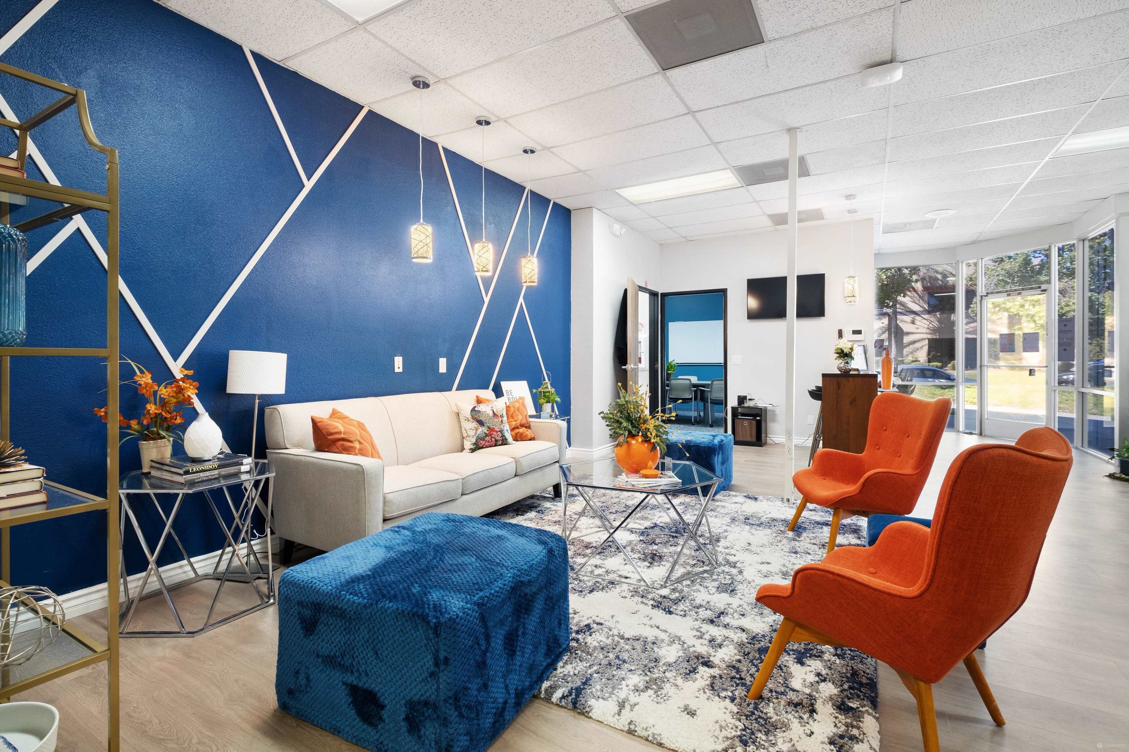A modern interior space with a light sofa, orange armchairs, a blue ottoman, and a geometric blue accent wall.
