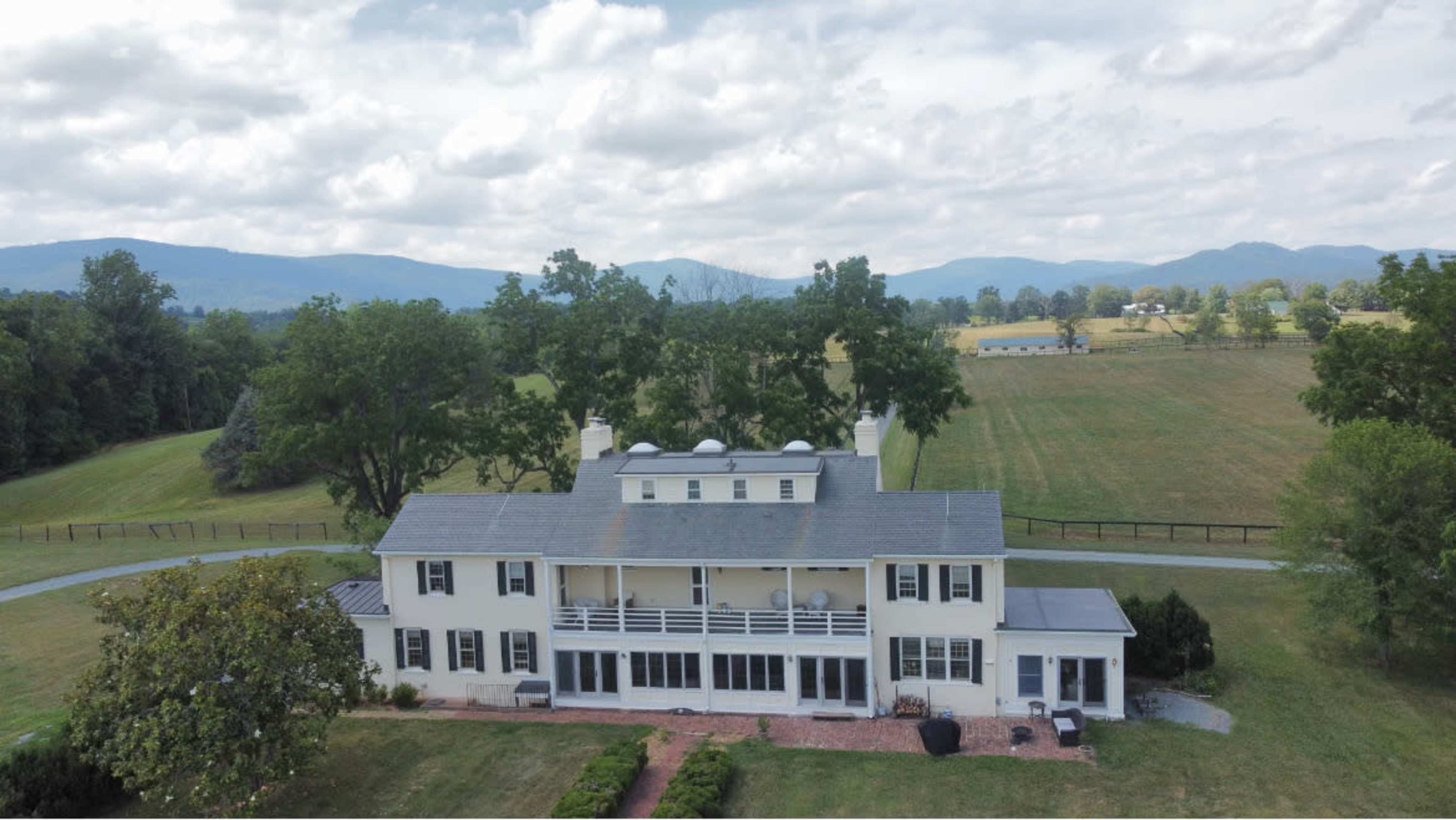 The image shows a large, white two-story house with a balcony and a patio, set against a backdrop of green fields and distant mountains under a cloudy sky.