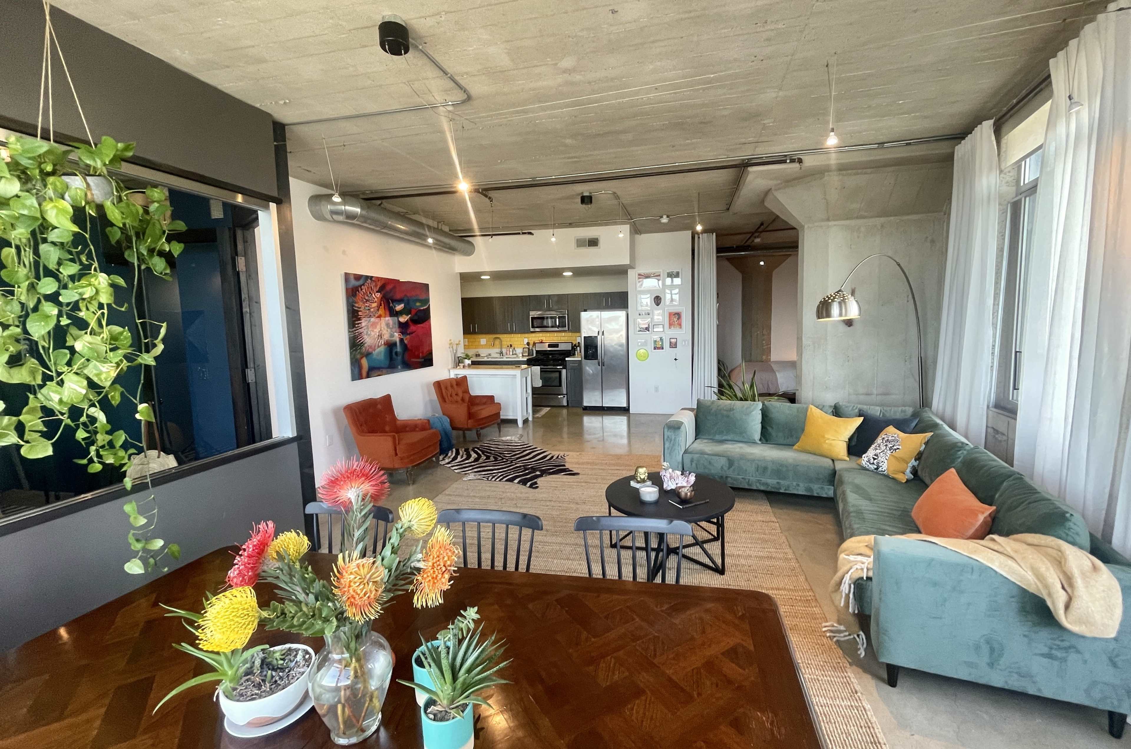 The image shows a modern open-concept living space featuring a green couch, two orange armchairs, a dining table with plants, and a kitchen area in the background.