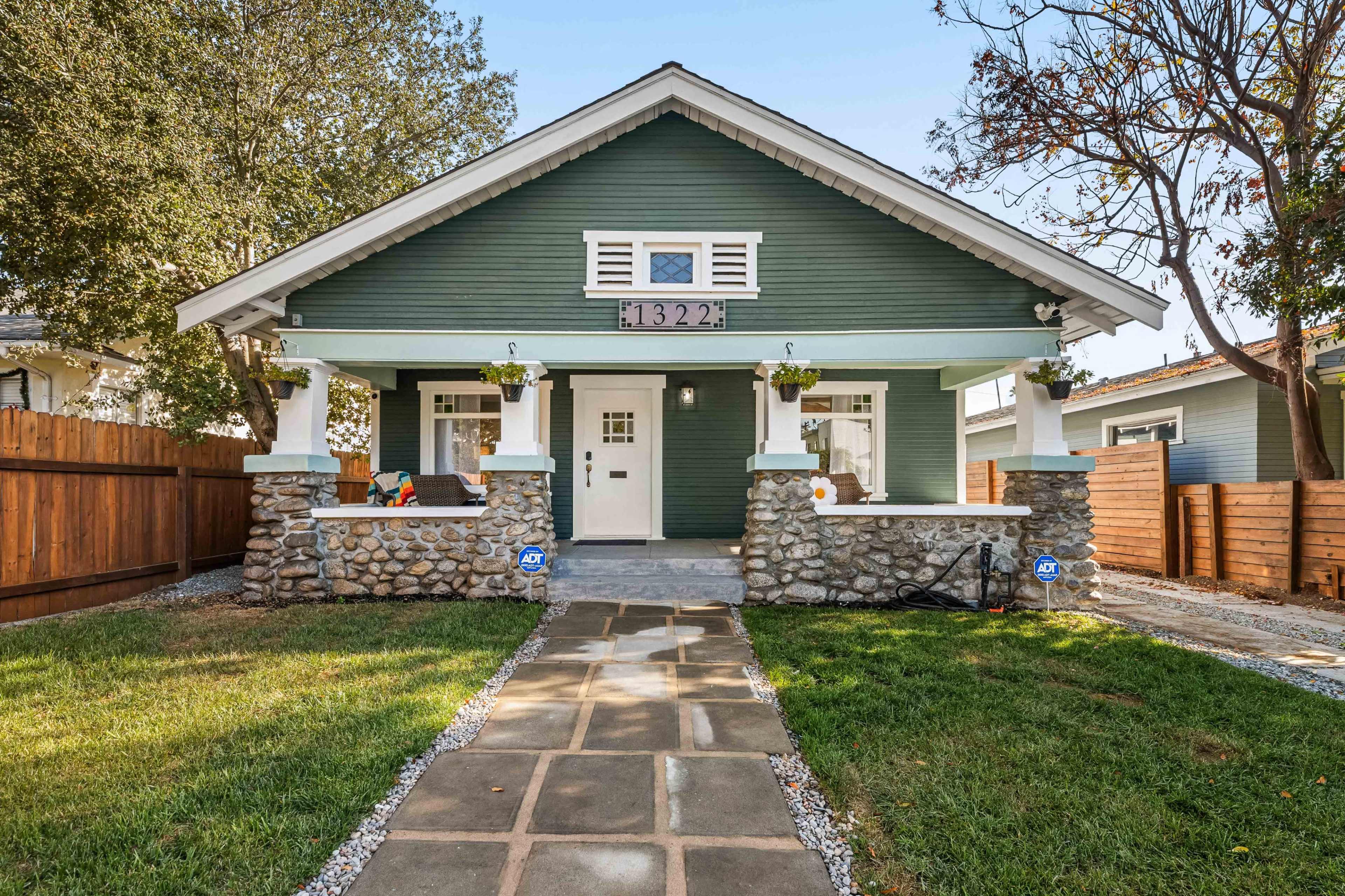 A single-story, green-painted house with stone accents and a front porch is framed by a grassy yard and paved walkway.
