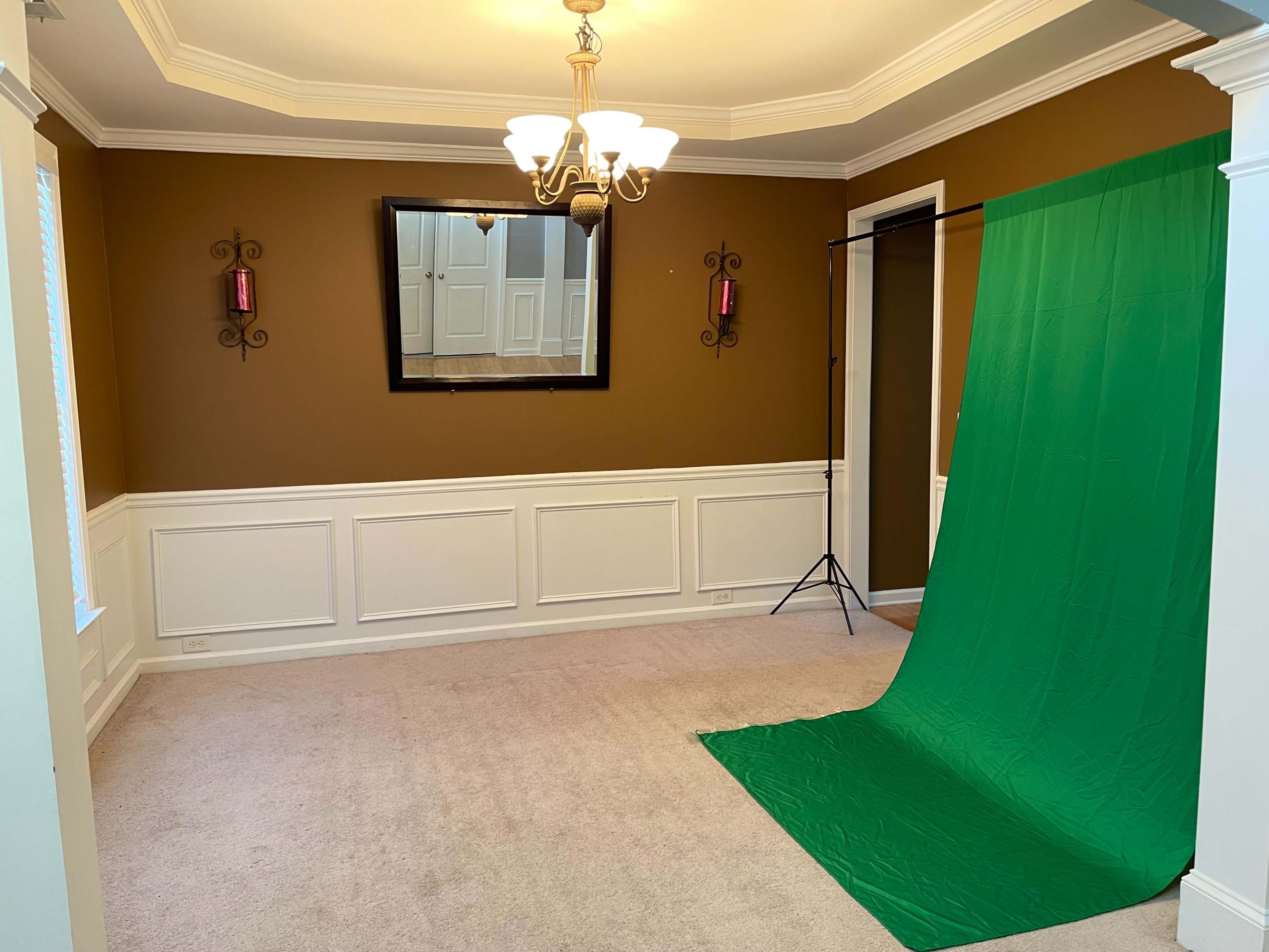 An empty room with brown walls, a chandelier, a large mirror, and a green backdrop set up in one corner.